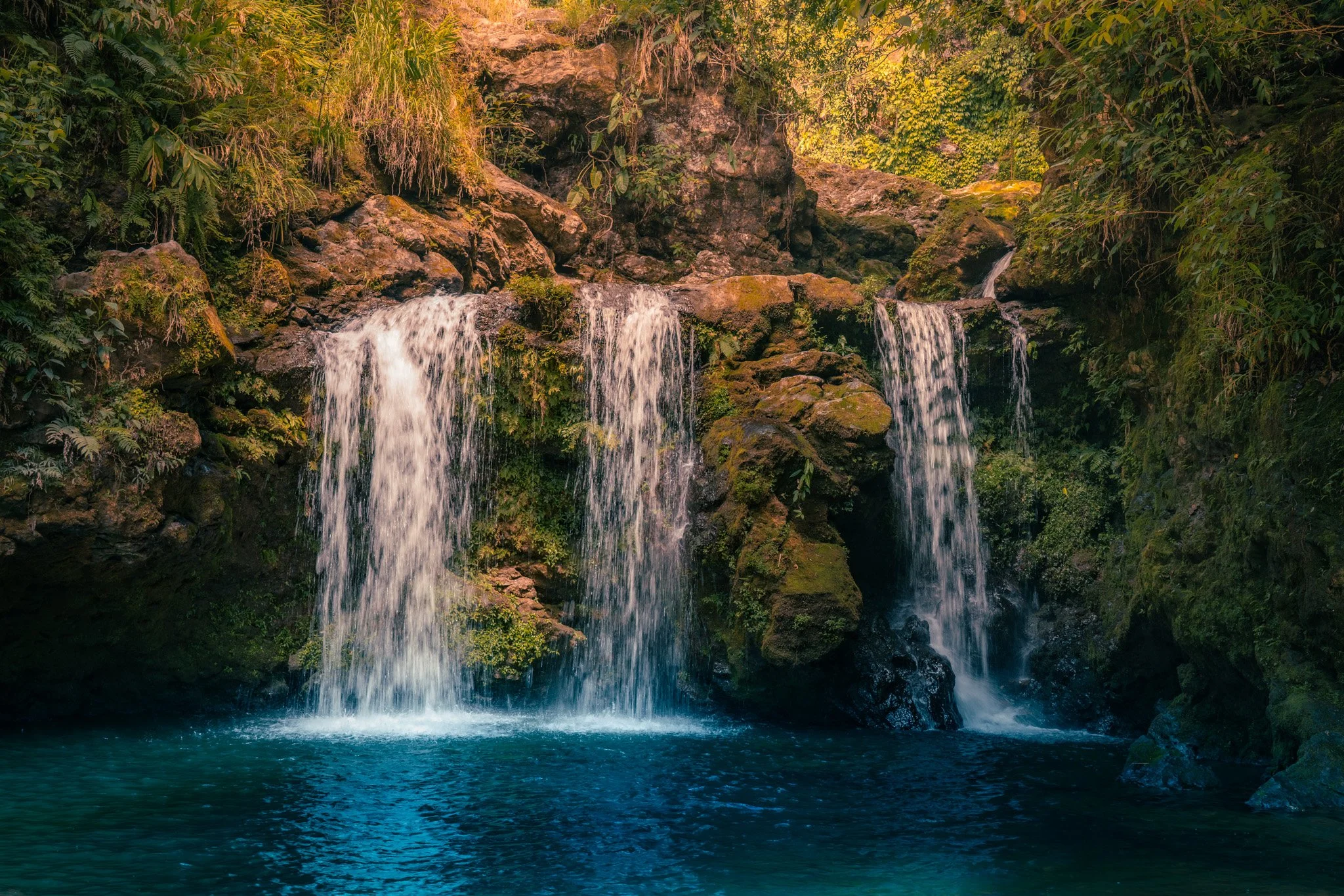 A serene waterfall flowing over rocks into a blue pool surrounded by lush green vegetation.