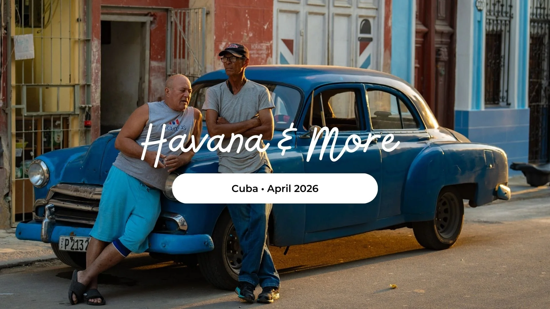 Two men conversing next to a vintage blue car on a street in Cuba, with colorful buildings in the background. One man is wearing a sleeveless shirt and shorts, the other is in a t-shirt and jeans, both are standing and leaning against the car.