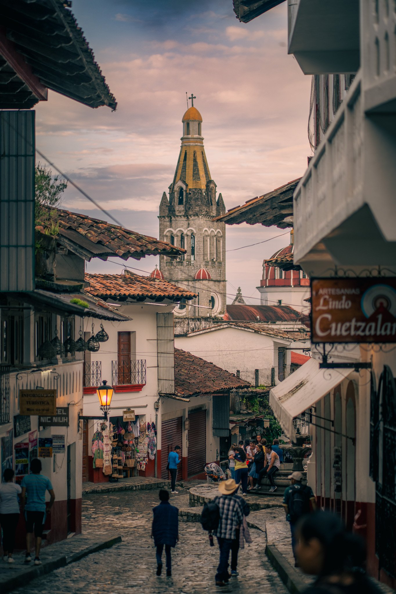 A cobblestone street in a historic neighborhood with shops and people walking, leading to a church tower with a cross on top, surrounded by old buildings with red-tiled roofs, during dusk.