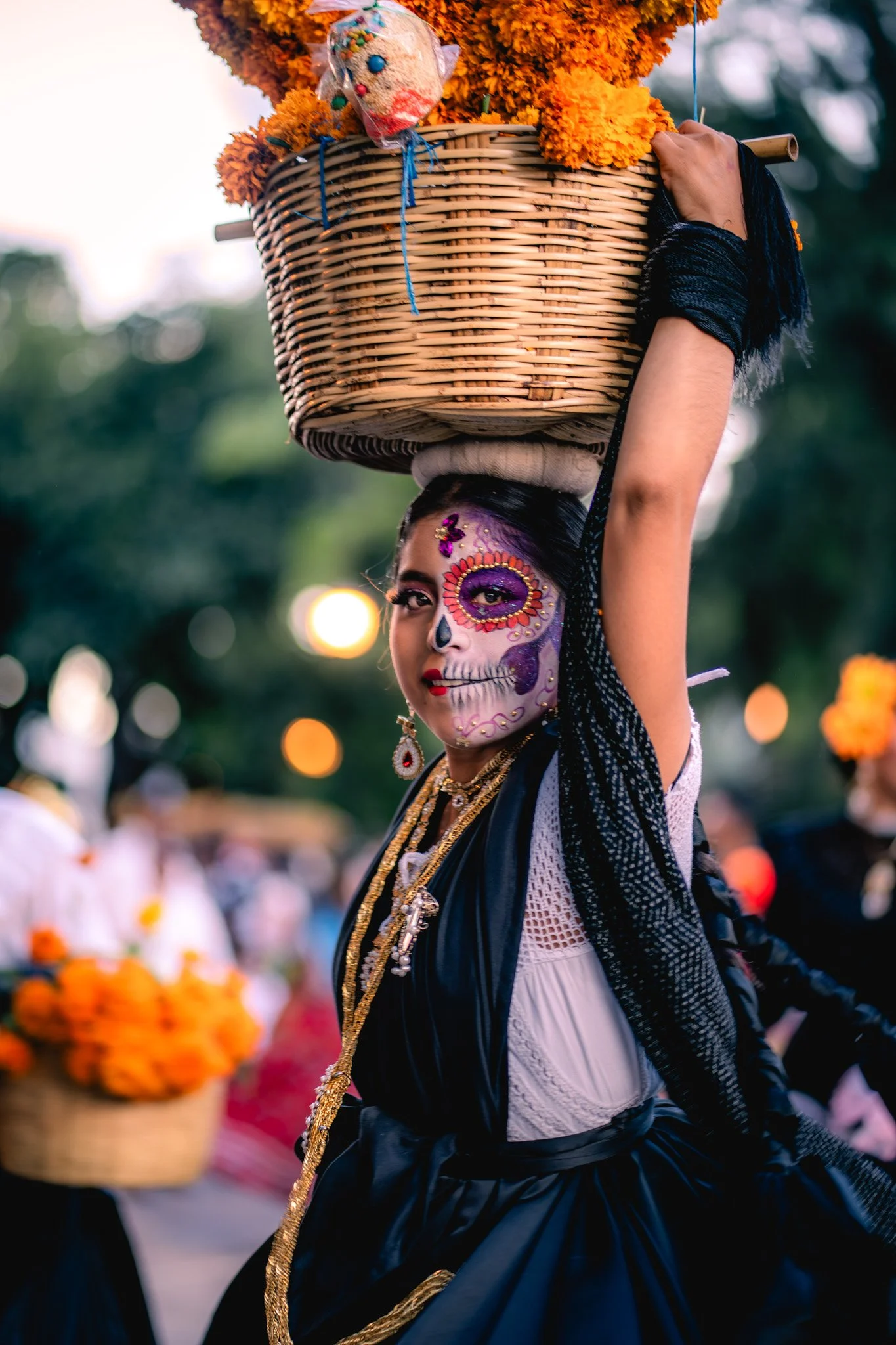 A woman participating in a cultural celebration, with her face painted in a colorful sugar skull design, wearing traditional attire, and balancing a basket of marigolds and a doll on her head.