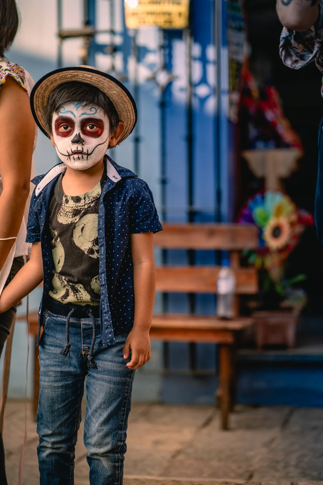 A young boy wearing face paint resembling a sugar skull, with black around his eyes, nose, and mouth, and blue decorative designs on his forehead, standing outdoors at a festive event.