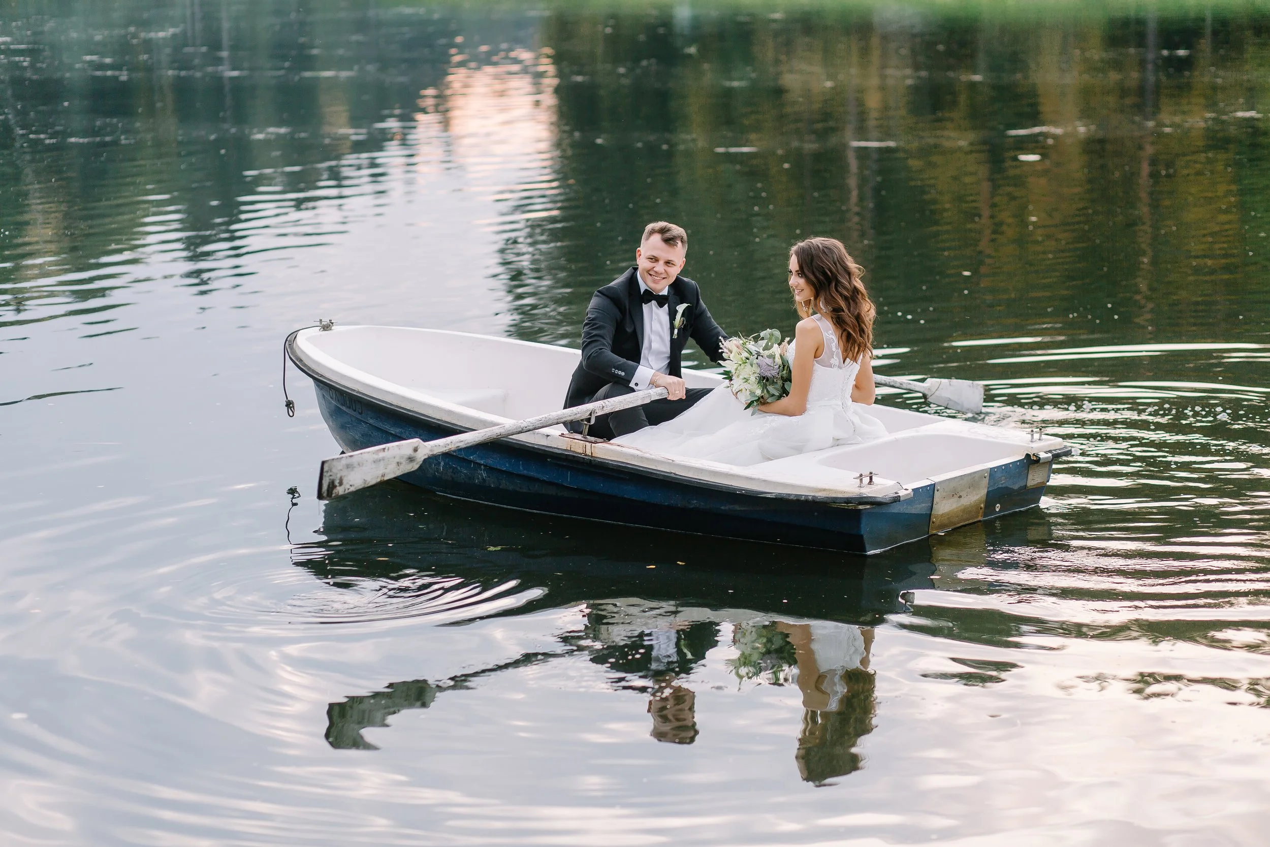 Bride and groom floating away on a row boat on lake. Bride is holding a bouquet of flowers and they are laughing.