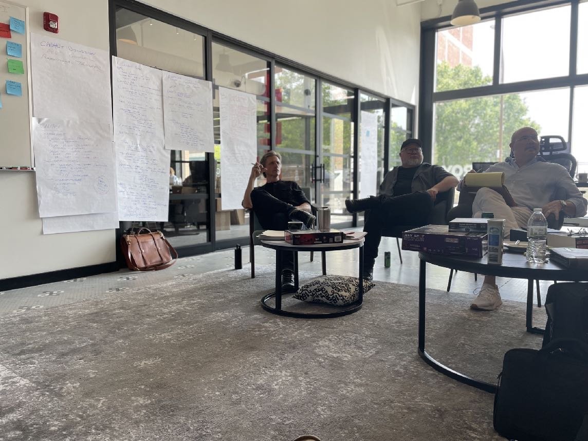 Three people sitting in a modern office with large windows and gray carpet. They are surrounded by tables with papers, books, and water bottles. White papers with writing are taped to the wall. A brown leather bag is on the floor.