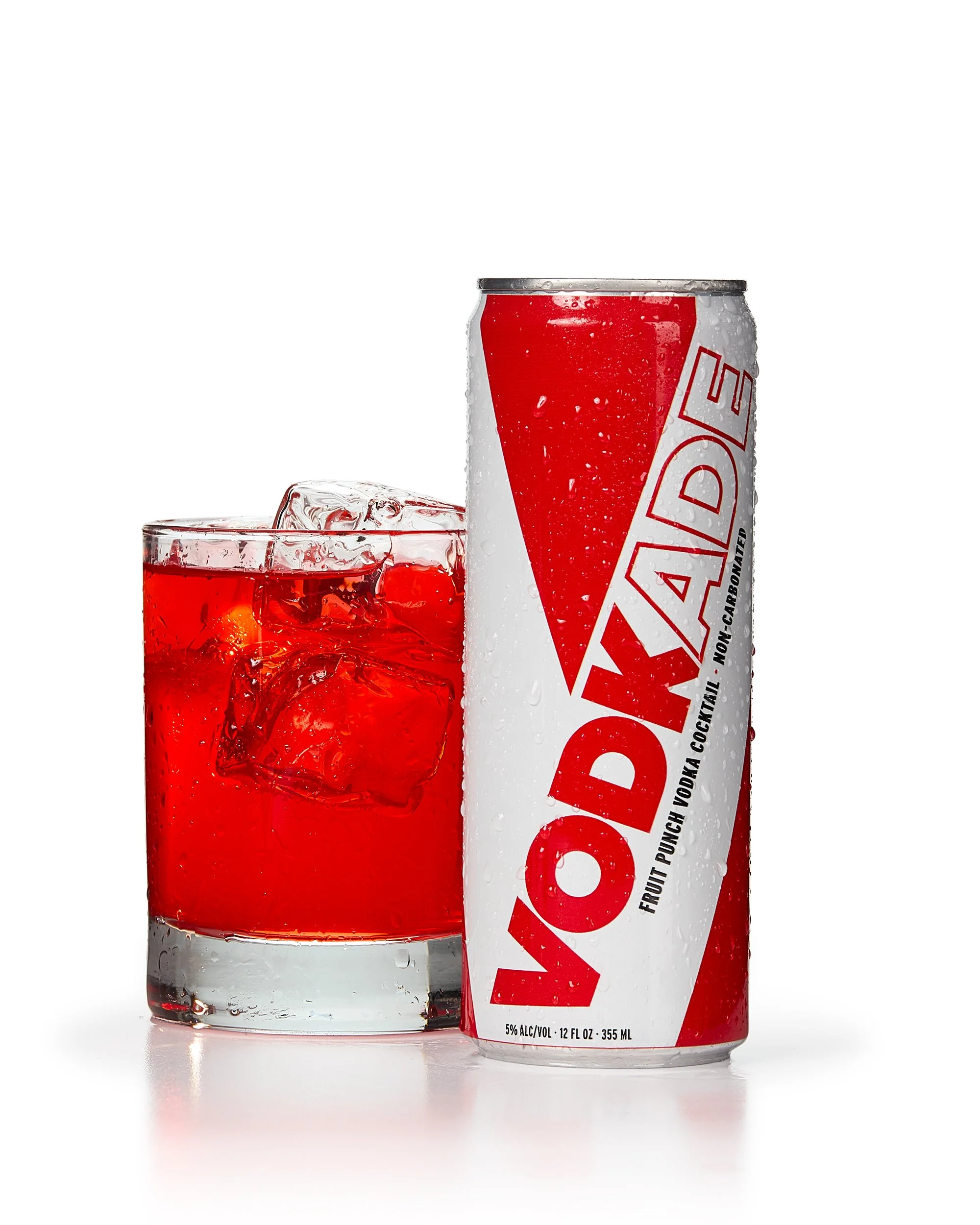 A can of VODKADE fruit punch vodka cocktail next to a glass filled with ice cubes and red liquid, both on a white background.