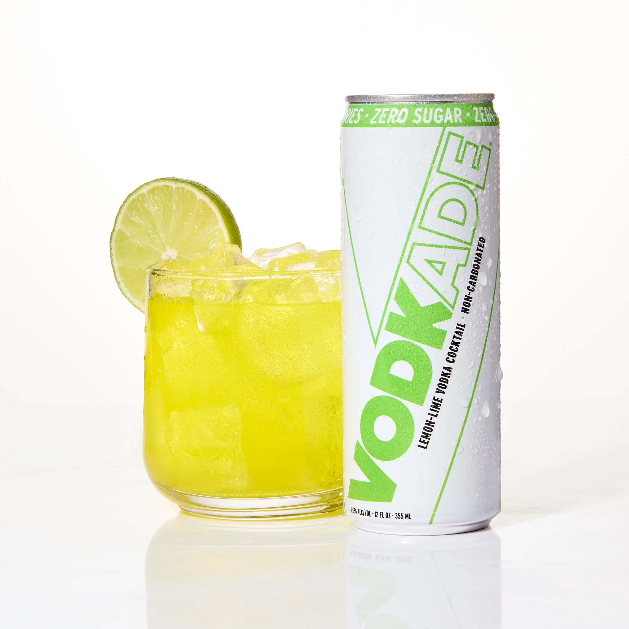 A can of lime-flavored Vodkade with condensation droplets next to a glass filled with lime Vodkade ice, and a lime wedge, on a white background.