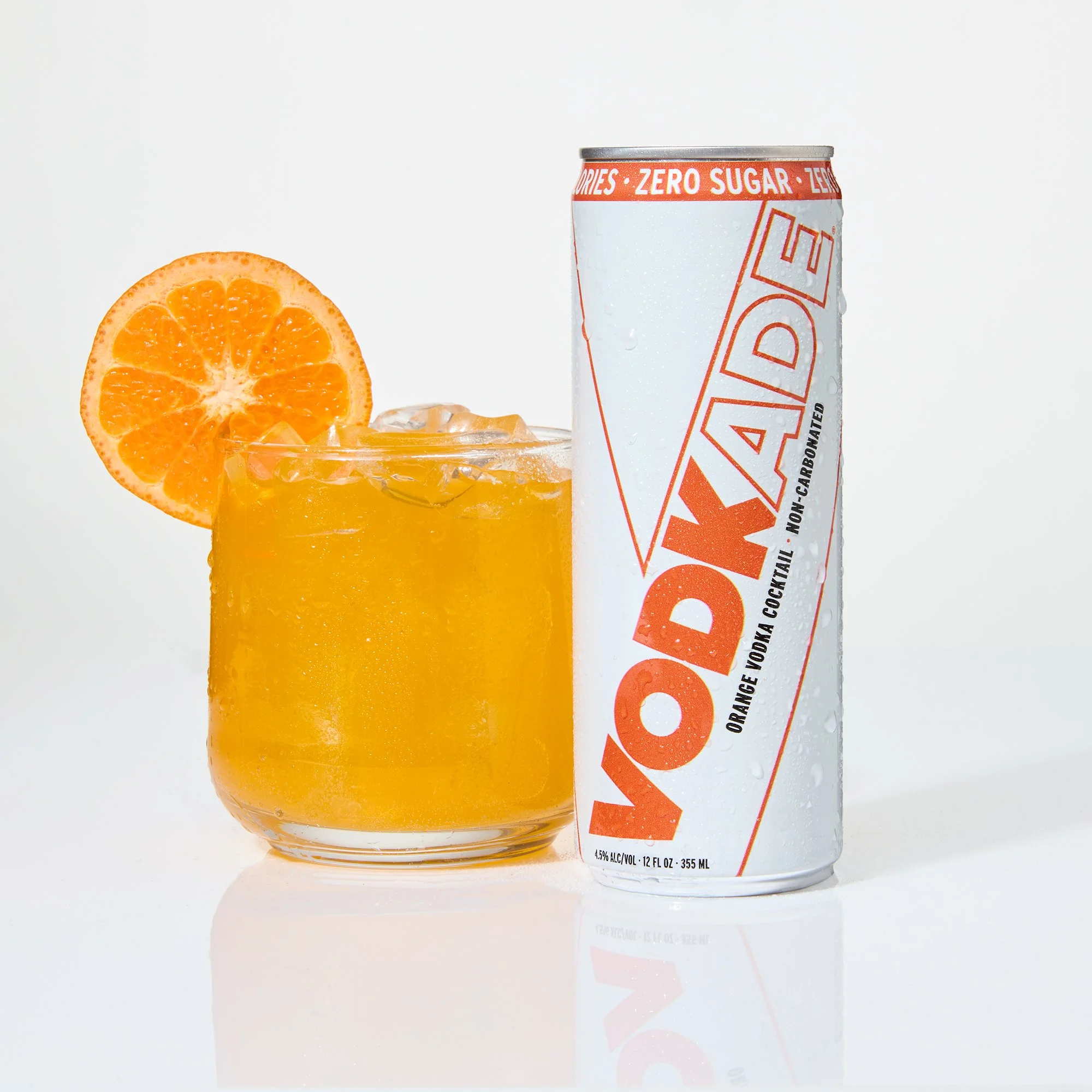 A glass of orange Vodkade with ice and a slice of orange on the rim, next to a can of vodkade with zero sugar on a white background.