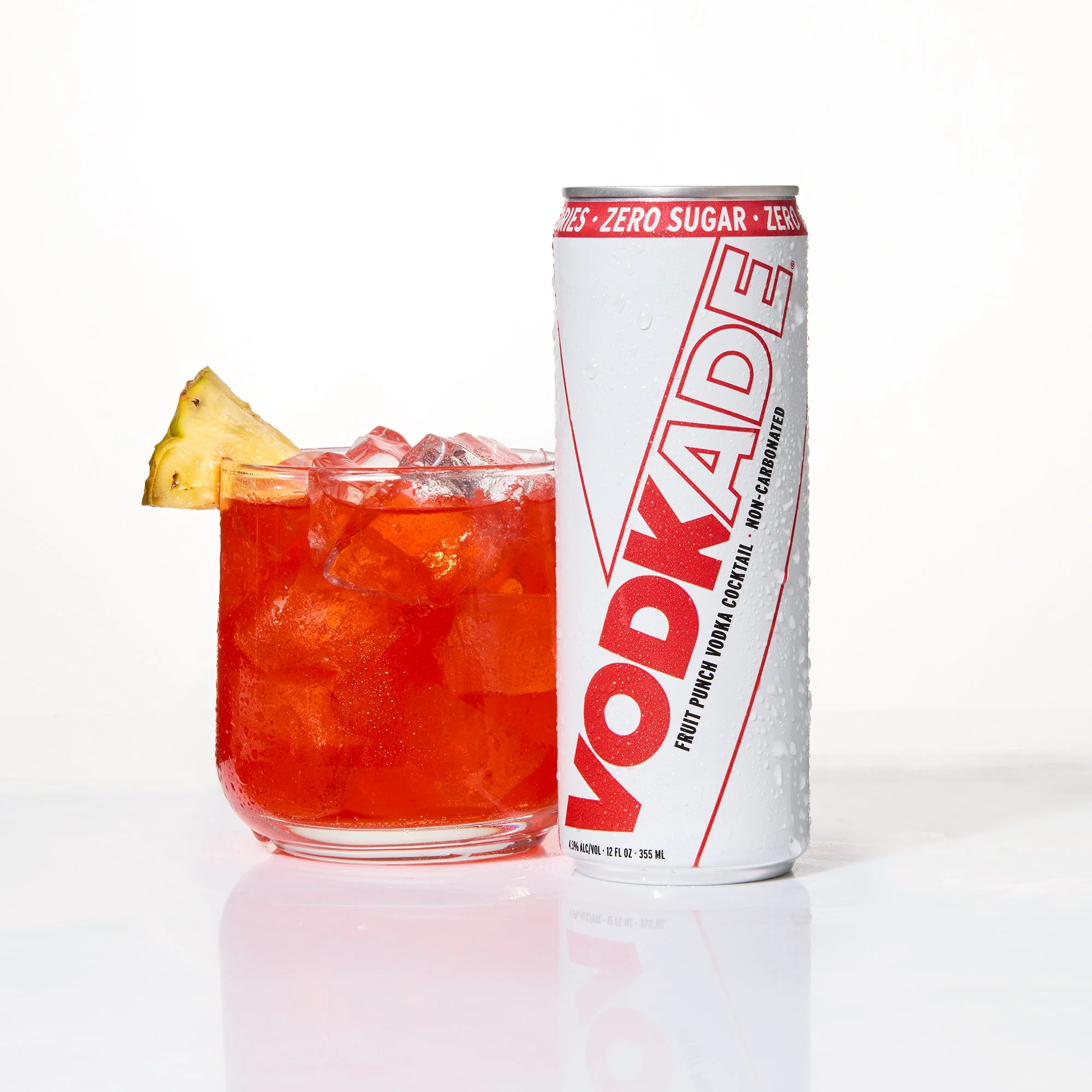A can of Vodkade zero sugar next to a glass filled with red liquid and ice, garnished with a pineapple wedge, against a white background.