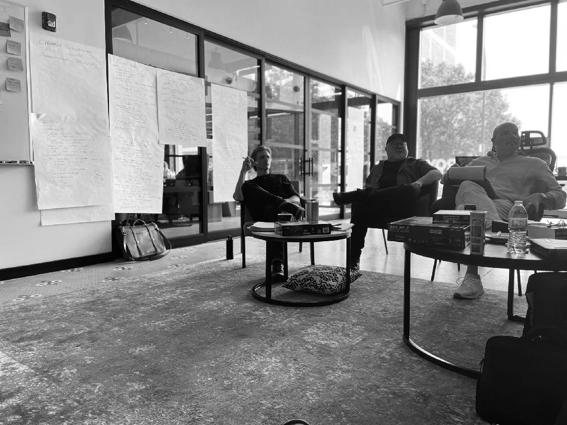 Three people sitting in a modern office with large windows taking part in a DESTROY DAY workshop, facing large sheets of paper filled with handwritten notes taped to the glass wall.