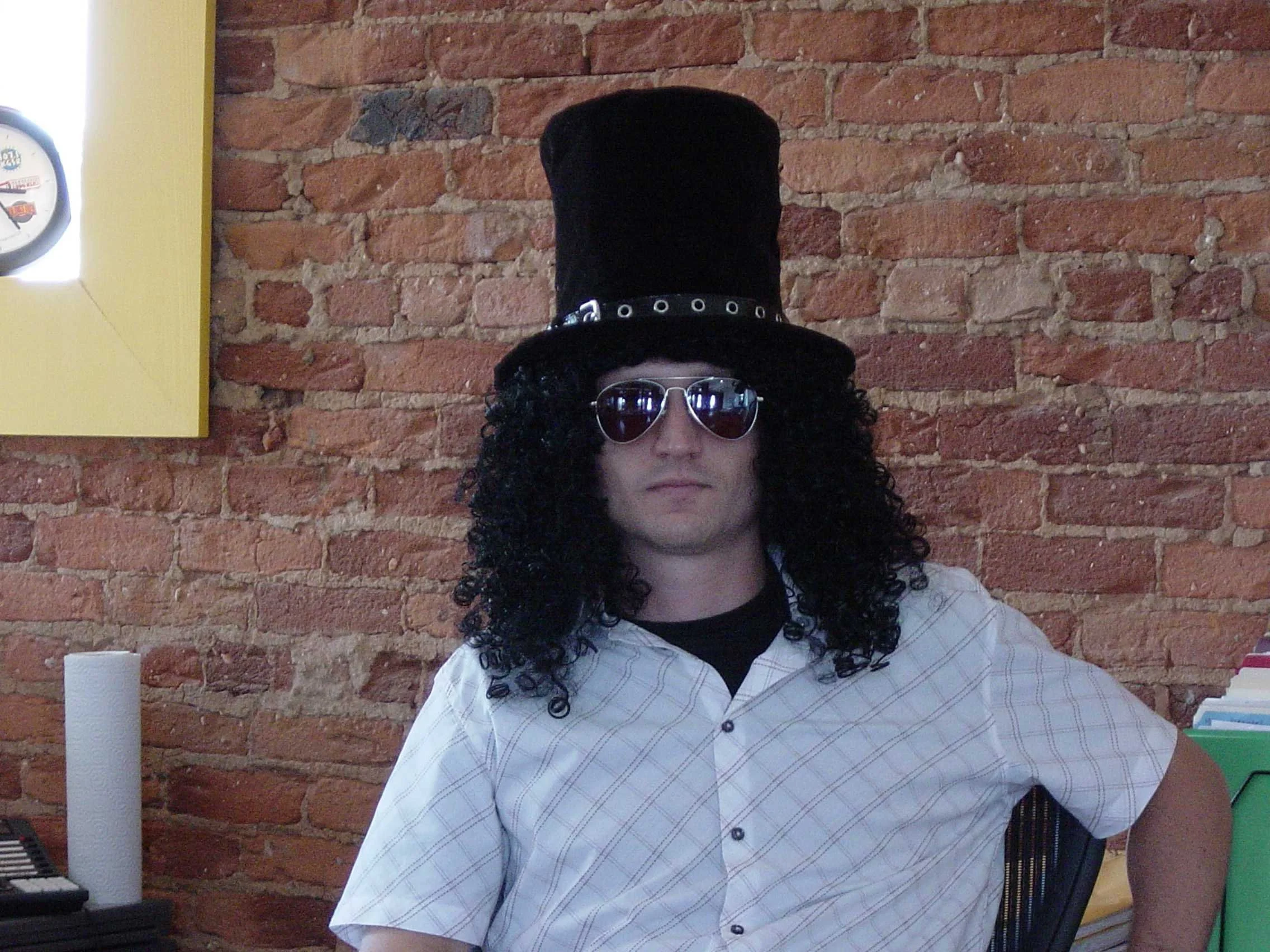 Person wearing a black top hat, curly wig, and sunglasses, sitting against a brick wall.