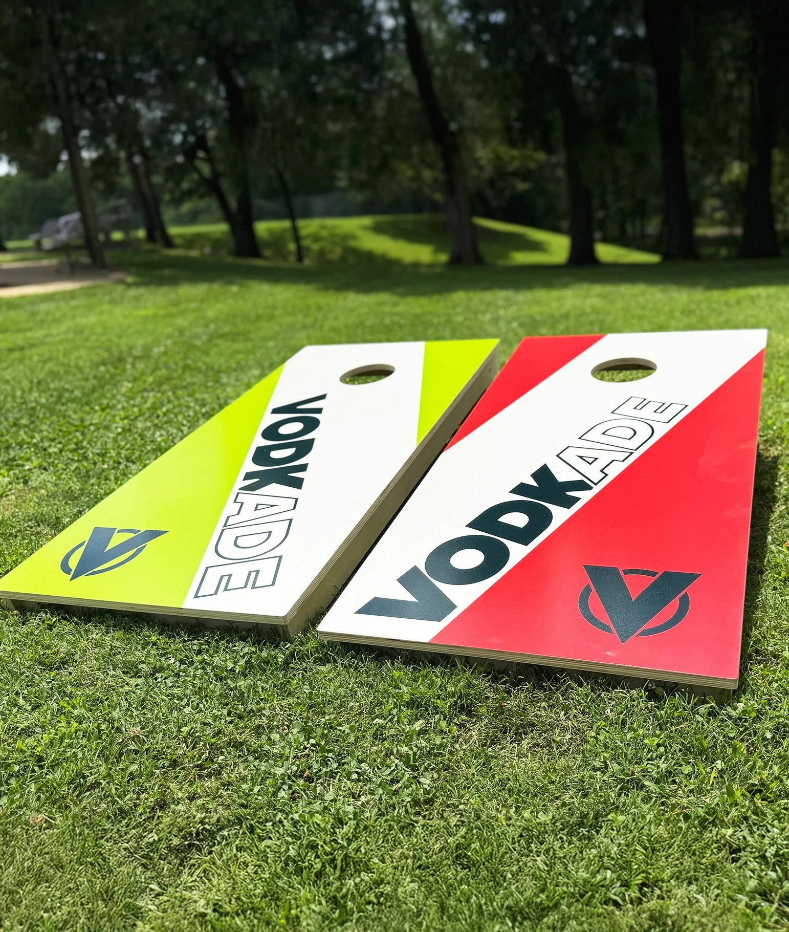 Two cornhole boards lying on a grassy lawn with trees in the background. One board is yellow-green and the other is red and white, both with the word 'VODKADE' and a logo.