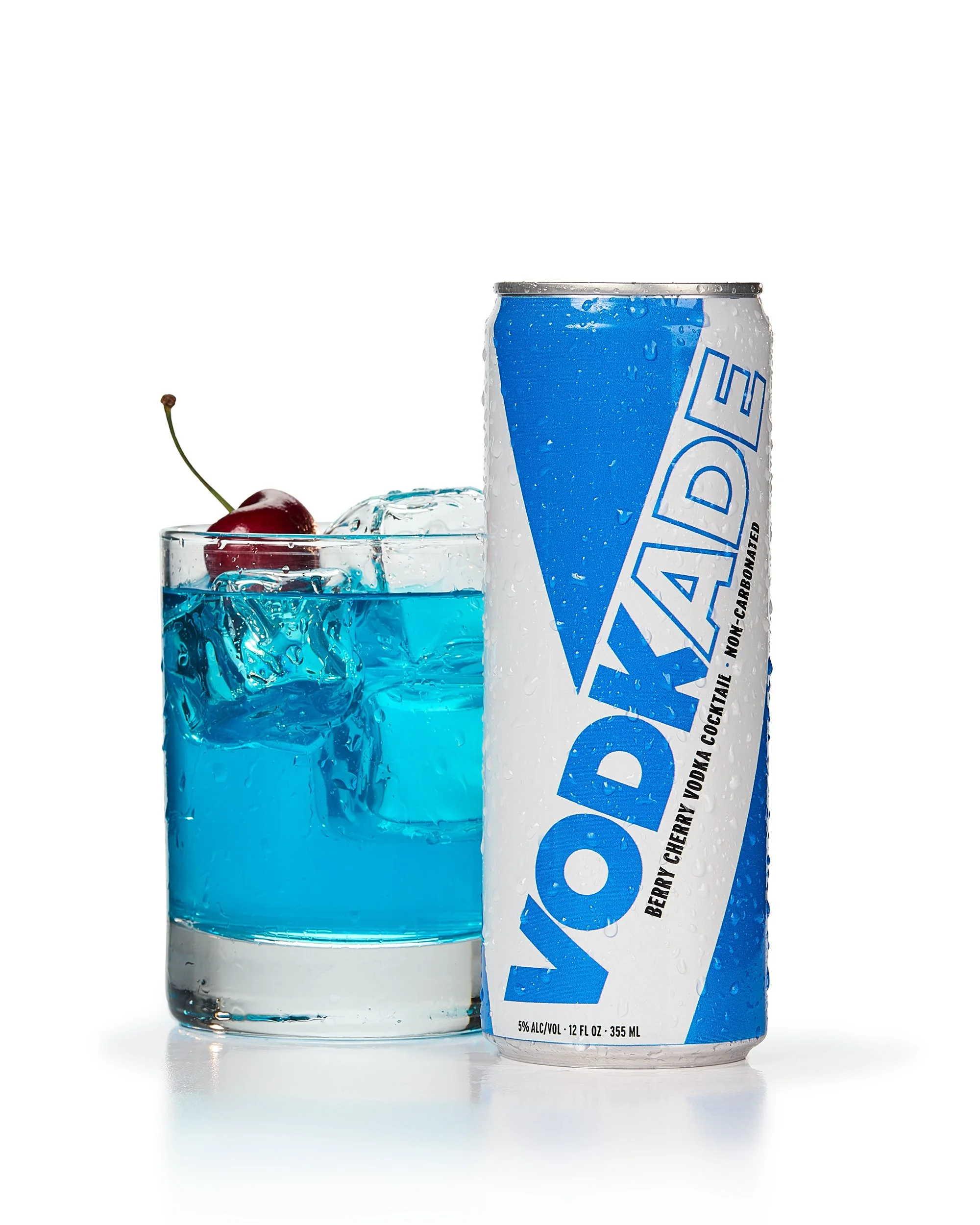 A blue can labeled "VODKADE" next to a glass of blue cocktail with ice cubes and a cherry garnish.