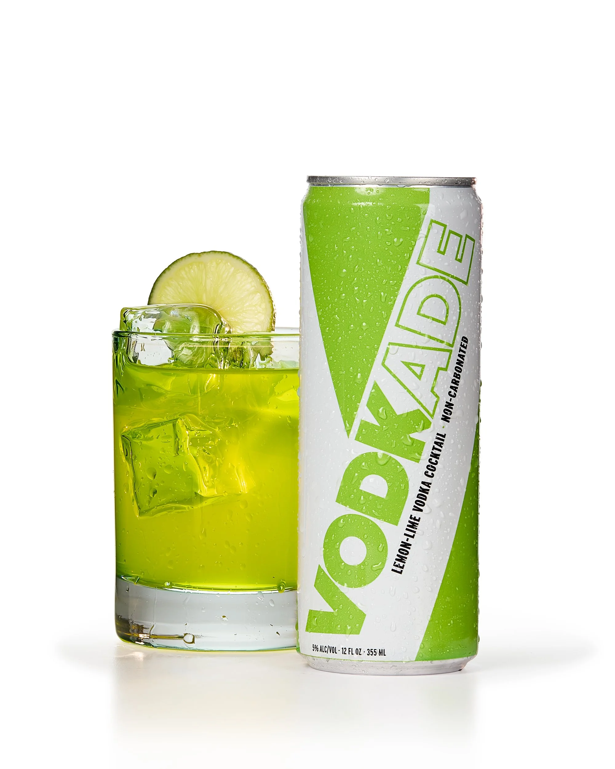 A can of Vodkade lemon-lime vodka cocktail next to a glass filled with green liquid, ice cubes, and a lime slice on the rim.