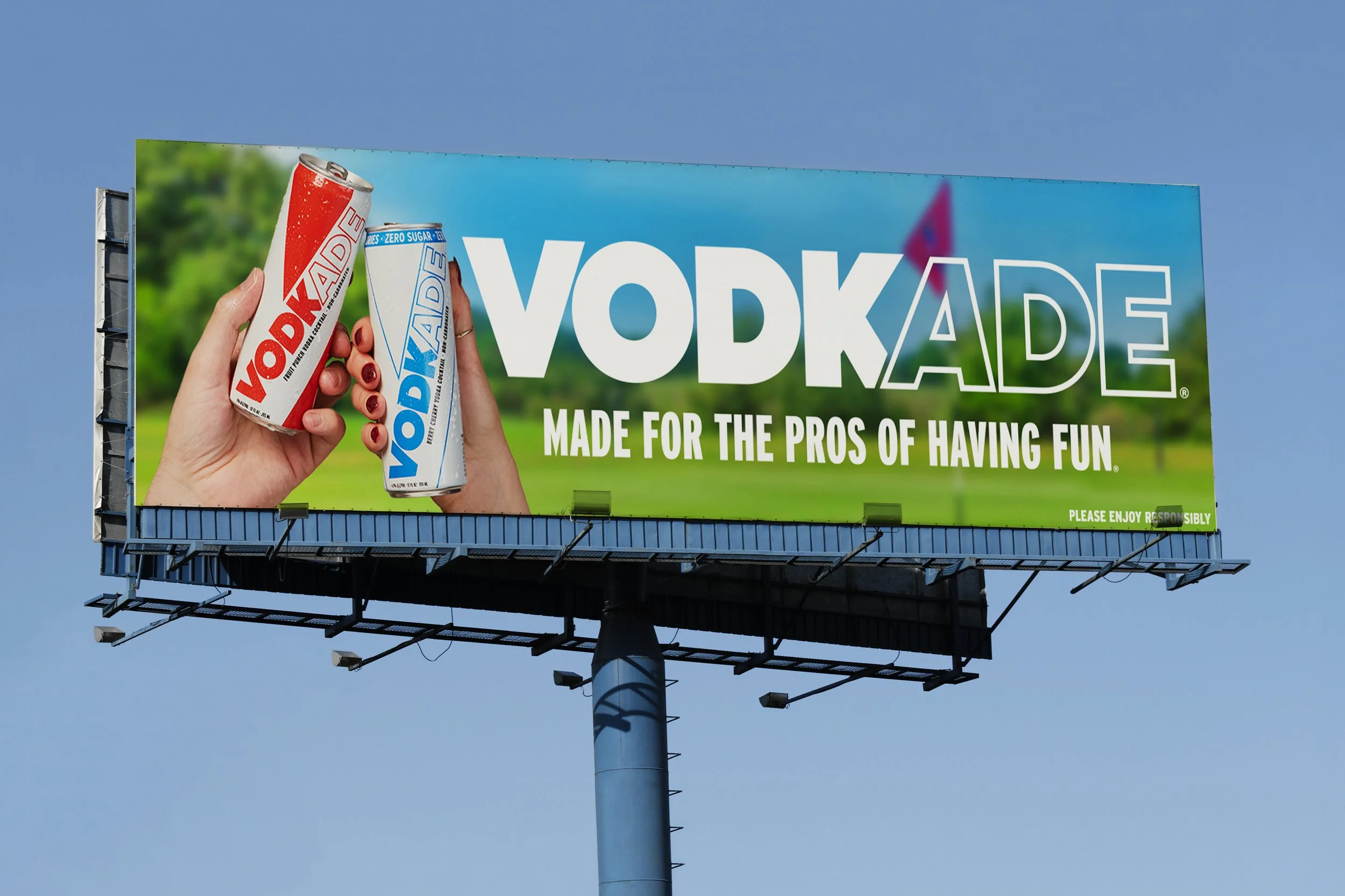 A billboard advertising Voodkade featuring two hands holding cans, one red and white and the other blue and white, against a park background. The billboard has large text reading "VODKADE" and a tagline "Made for the pros of having fun," with a legal