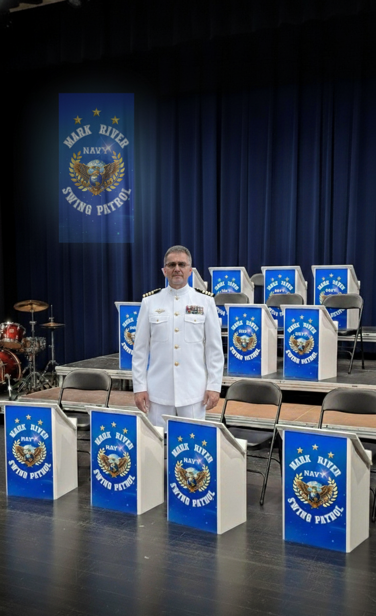 Een man in een witte marine-uniform staat voor een podium met stoelen en microfoons, versierd met borden en banners met het logo van de 'Mark River Navy Swing Patrol'. Op de achtergrond een blauwe gordijn en een drumbatter op de linkerzijde.