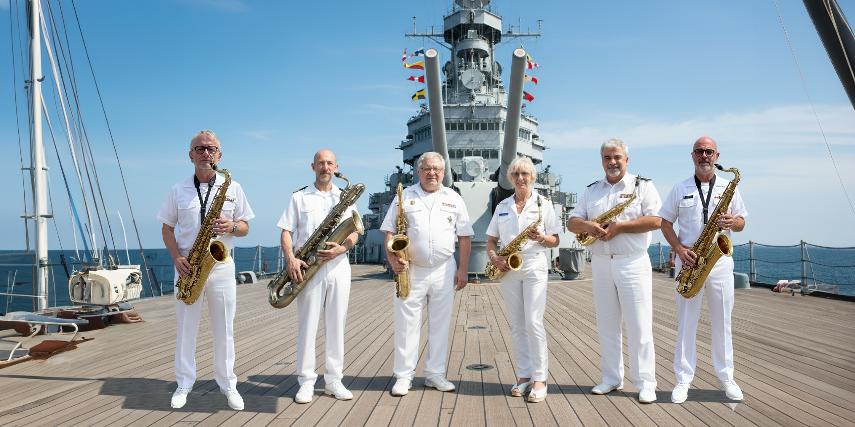 Een jazzband bestaande uit zes leden in witte uniformen met saxofonen, standing op het dek van een schip met een oorlogsschip op de achtergrond tijdens zonnig weer.