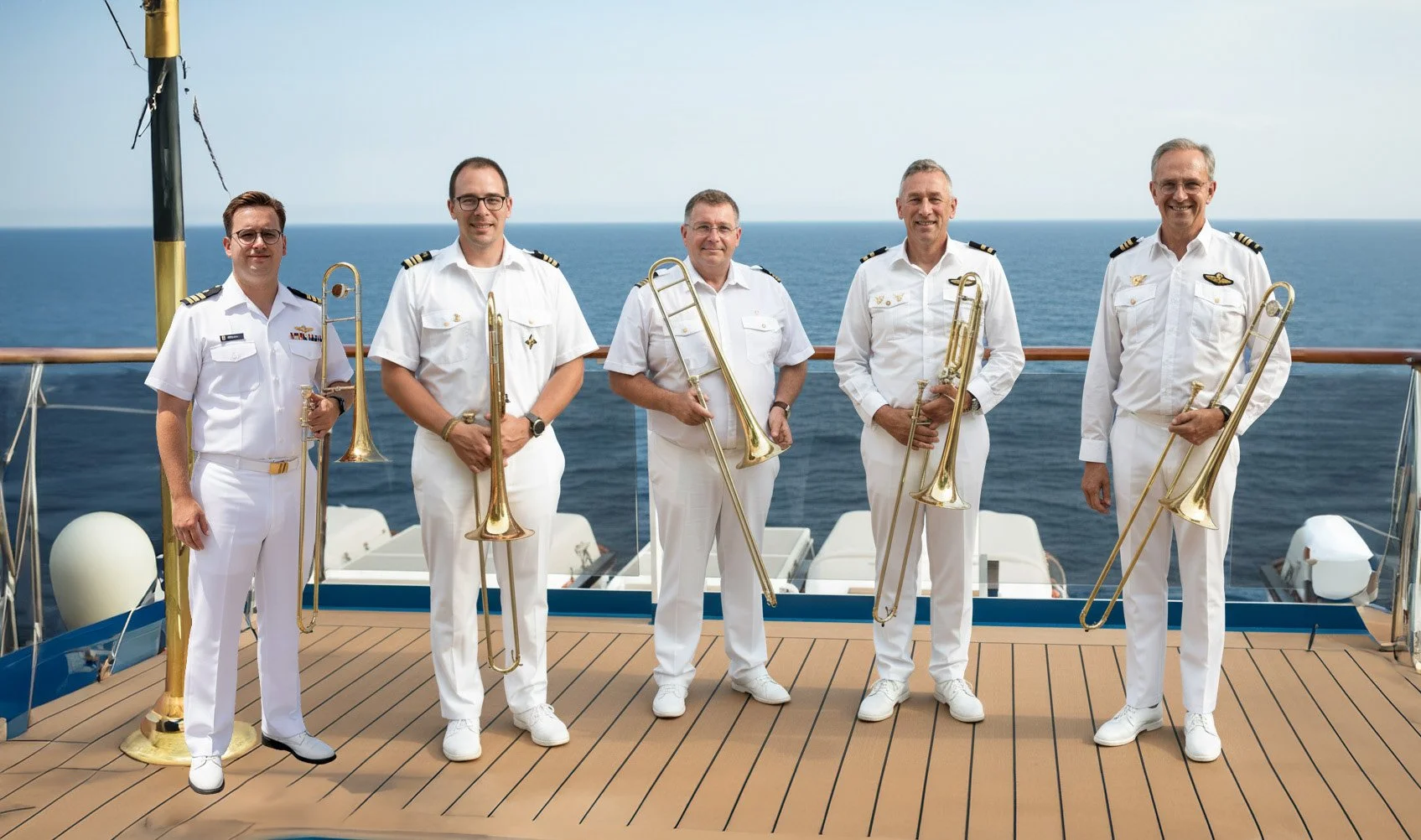 Vijf mannen in witte zeevaartuniformen die staan met een bi धनल और झुनझुना, op de vloer van een schip met de oceaan op de achtergrond.