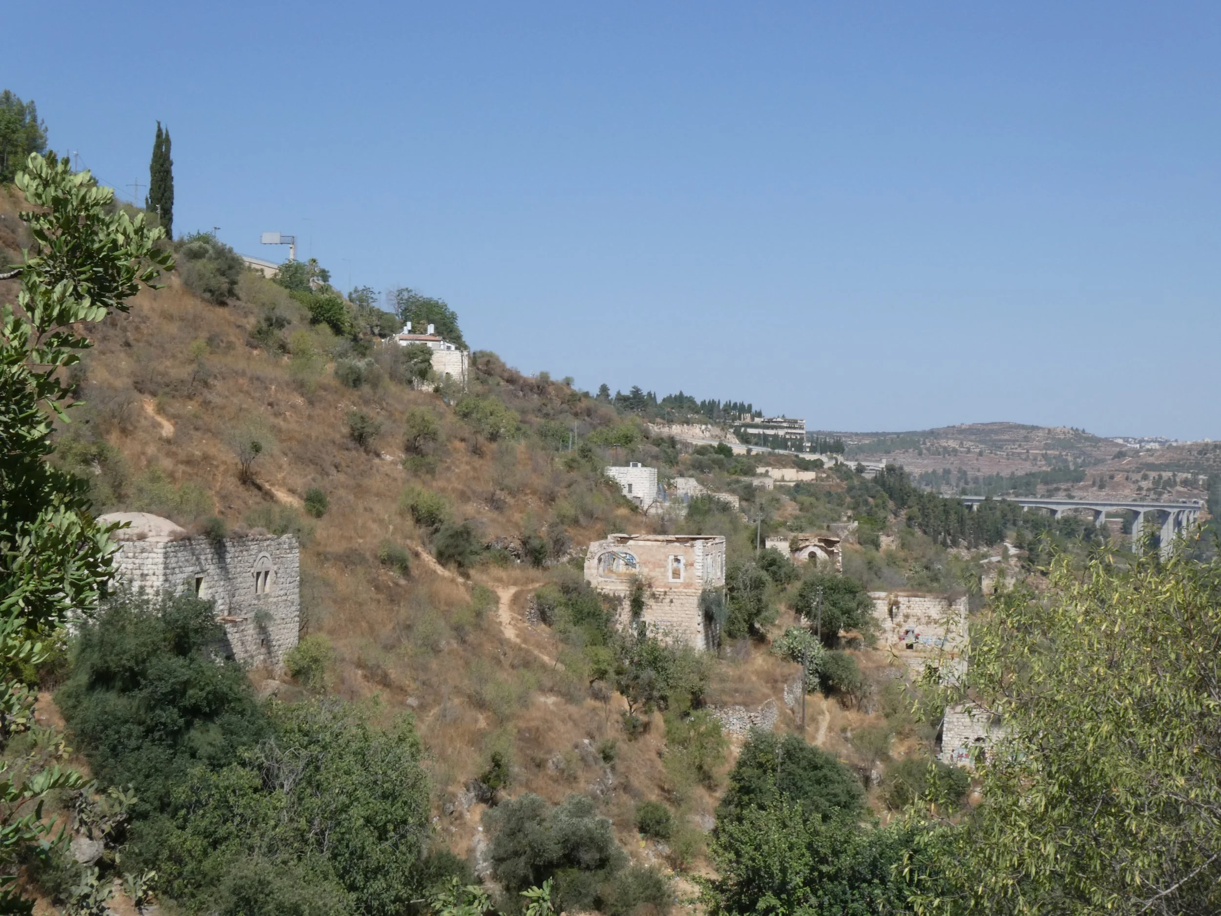 A view of a hillside with brown brush and green trees, along with several stone built houses which are missing their roofs.