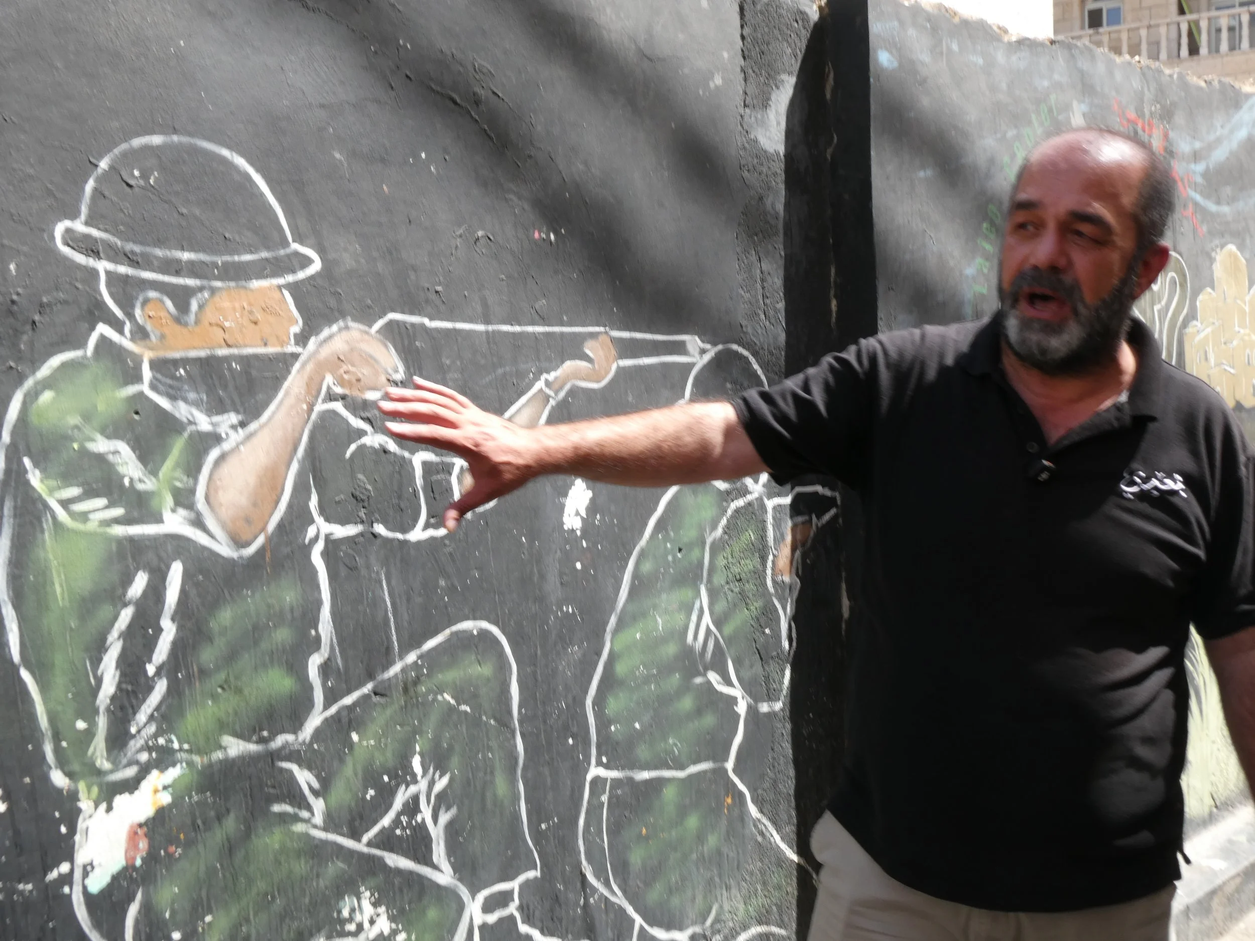 Palestinian man in black shirt pointing to a mural on a cement wall, which depicts a solider in green fatigues is seen kneeling and holding a rifle.