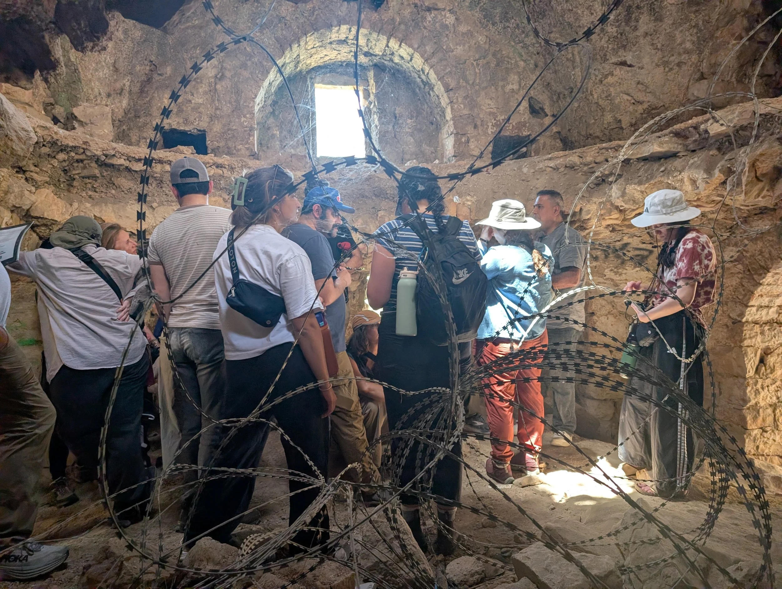 Eight people with hats, backpacks, and water bottles listening to a tour guide inside a stone building with an arch window. The forefront of the photo includes barbed wire.