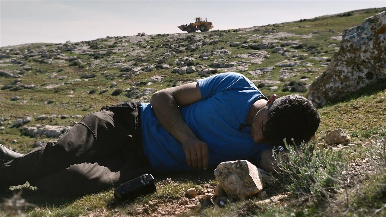 Promo picture of a Palestinian man with a camera lying on a hill with a bulldozer in the background.