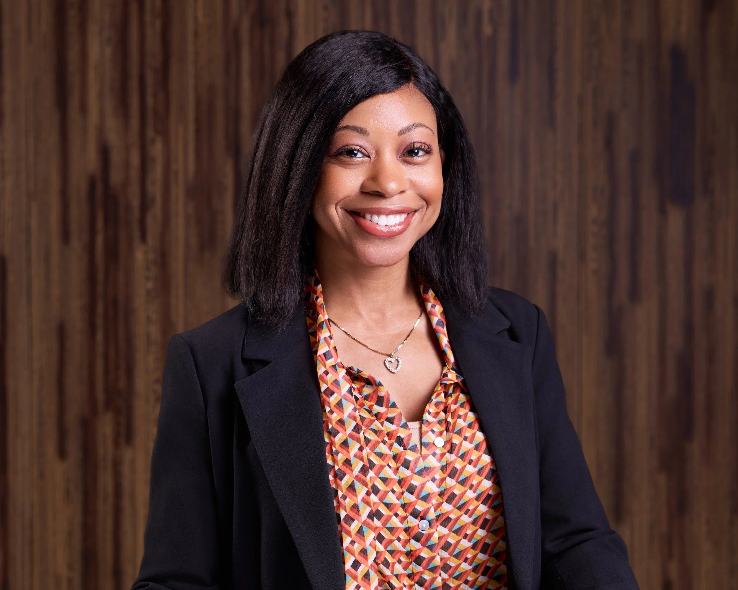 A woman with shoulder-length black hair smiling at the camera, dressed in a black blazer over a colorful patterned blouse, standing in front of a wooden background.