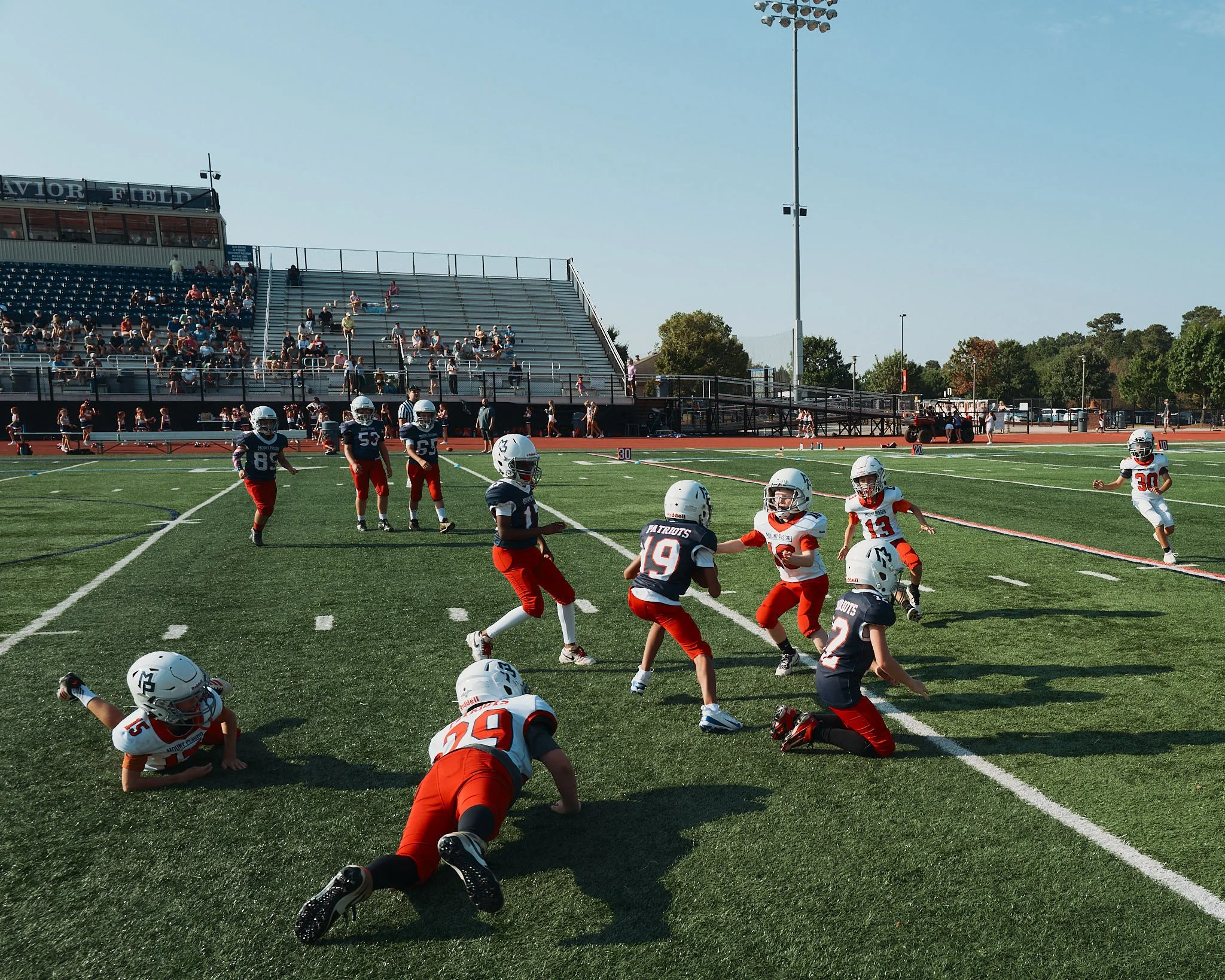 Young boys playing football on a field with spectators in the stands and trees in the background.