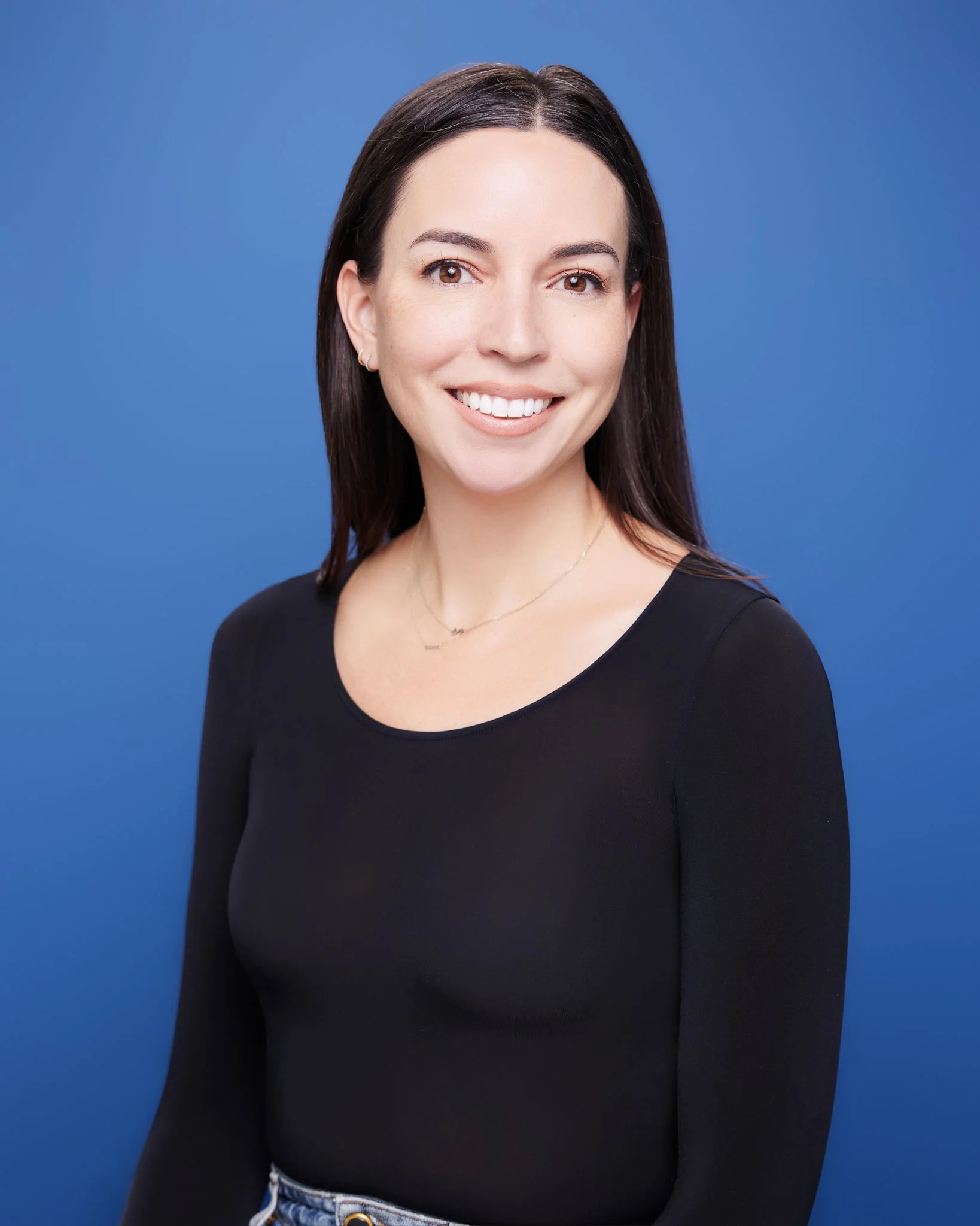 Portrait of a woman with dark brown hair, wearing a black top and a delicate necklace, smiling against a blue background.