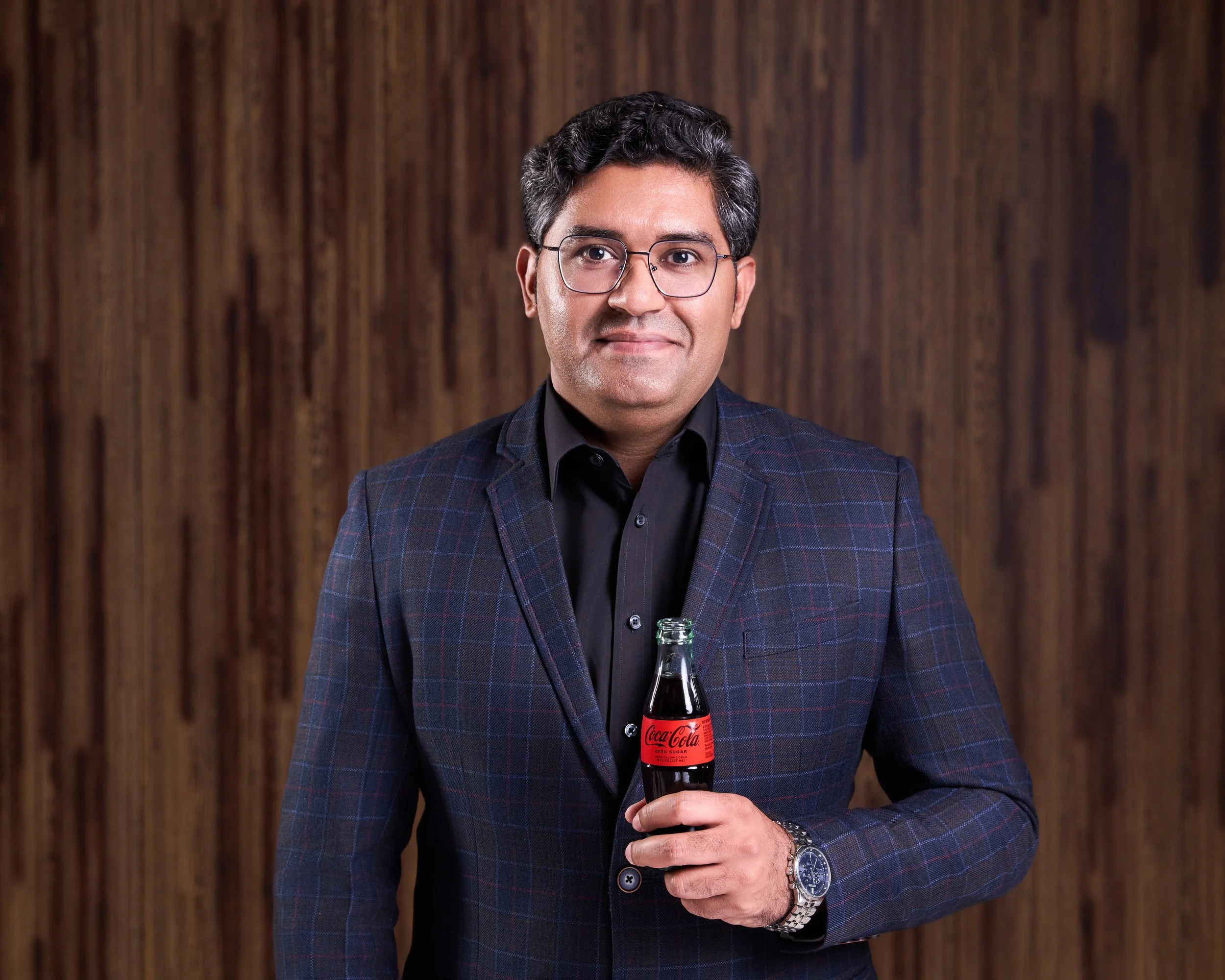 A headshot of a man with dark hair and glasses wearing a dark plaid blazer holding a Coca-Cola bottle, standing against a wooden background.