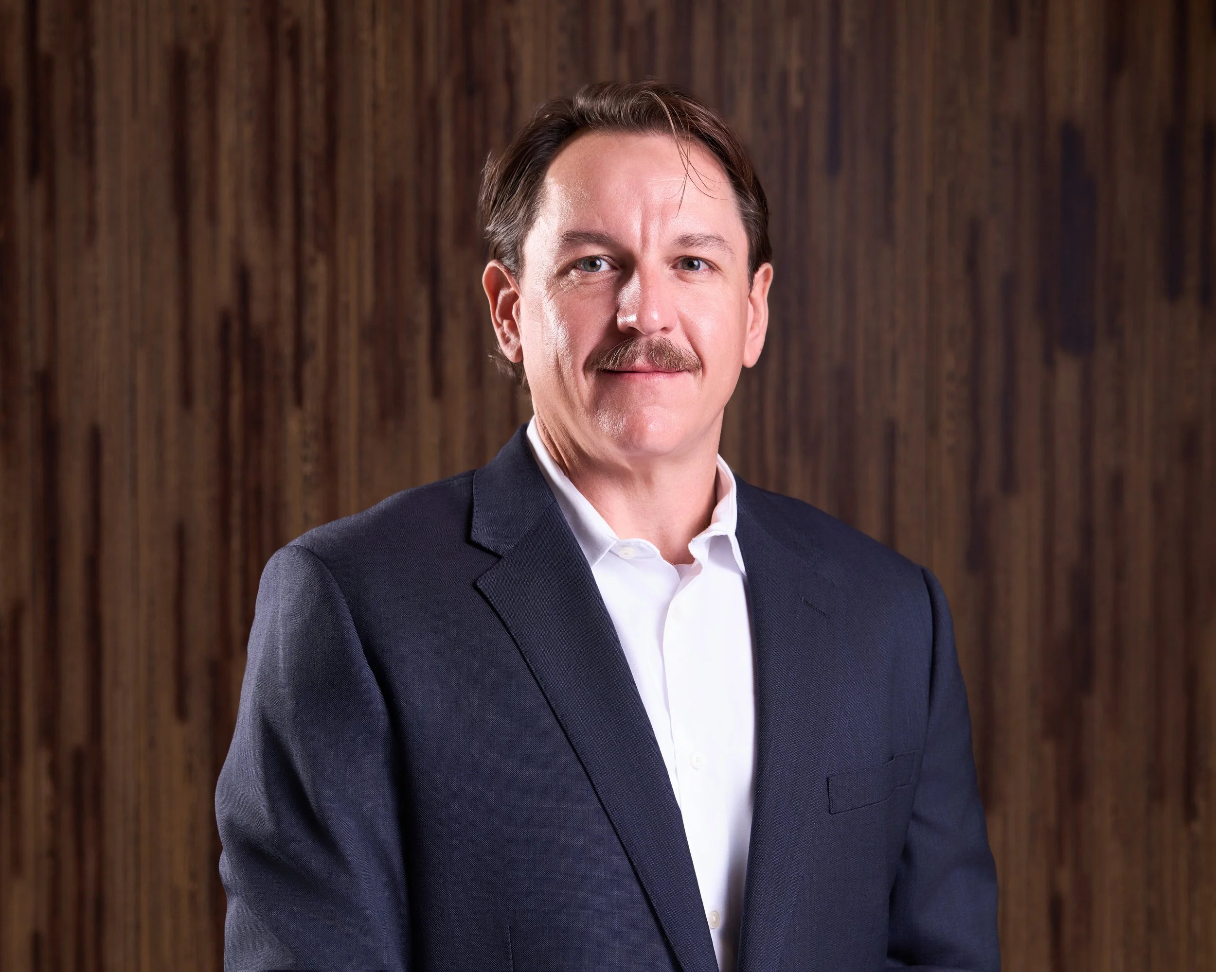 Professional headshot of a man with brown hair, wearing a dark suit and white shirt, standing against a wooden background.