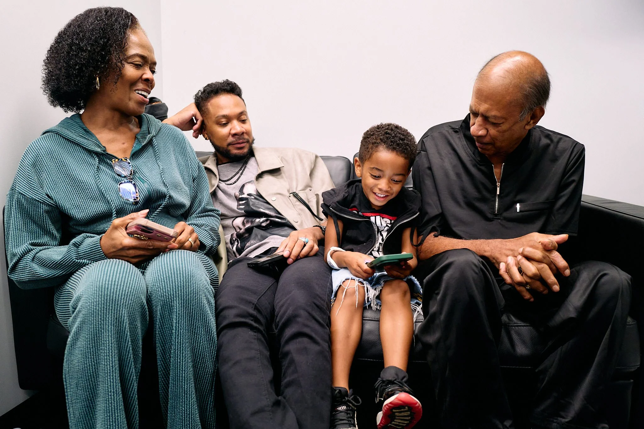 A multigenerational family of five sitting on a black leather sofa, using smartphones and engaging with each other in a casual setting.