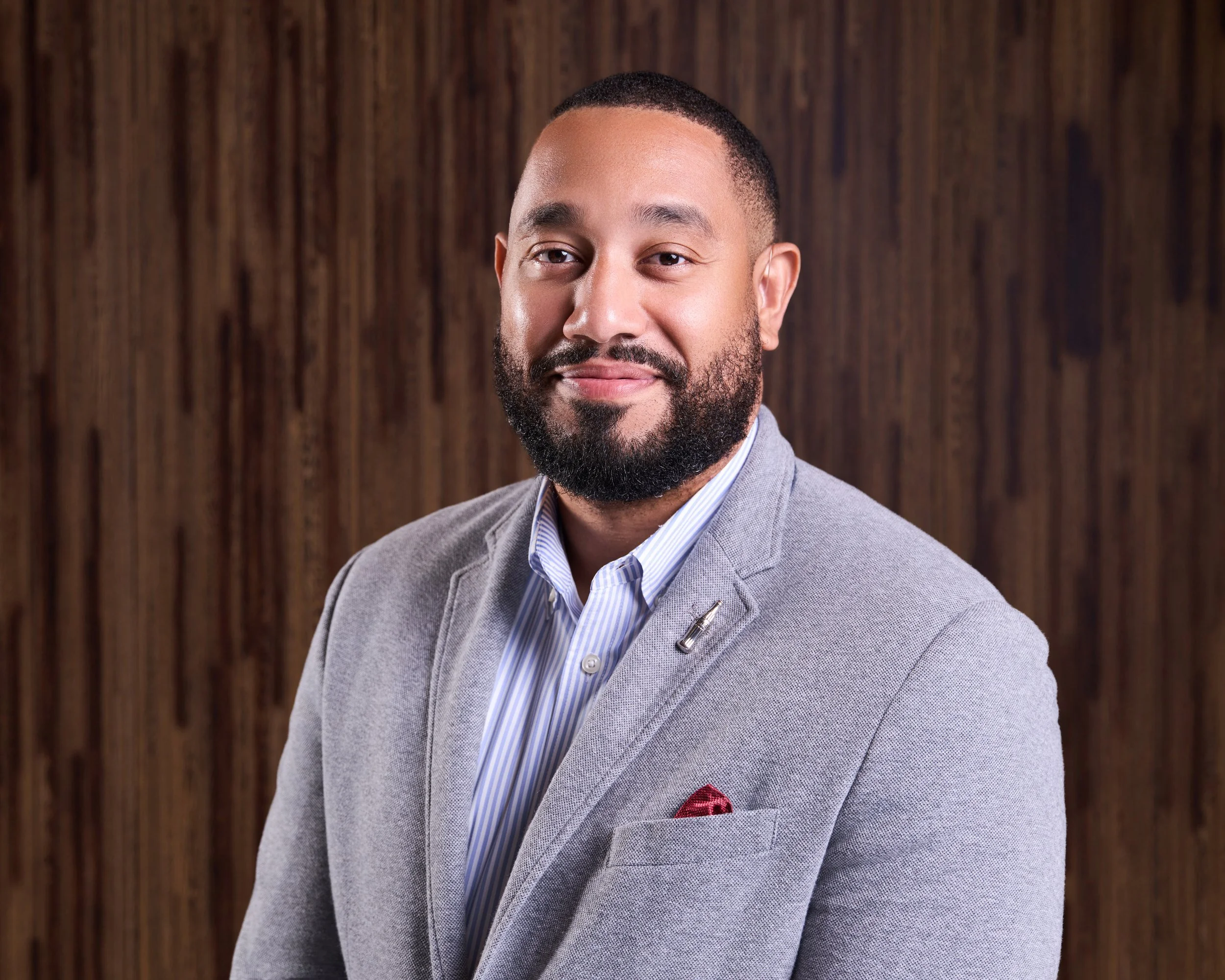 Headshot of a man with a beard wearing a light gray blazer, a striped shirt, and a red pocket square, smiling in front of a wooden background.