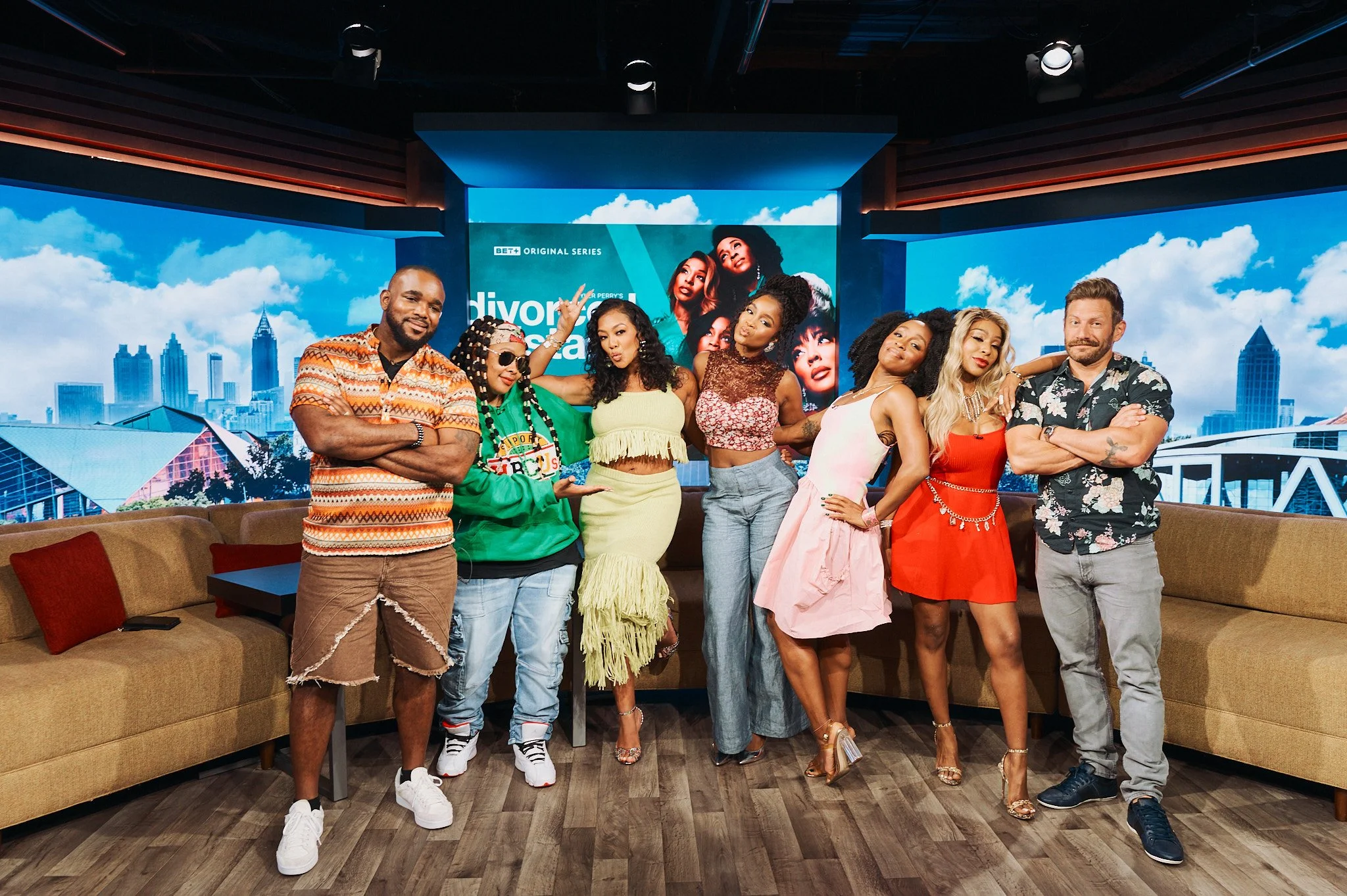 Khadeen Ellis and Cast are standing in a row in TV studio, posing for a photo in front of a large digital screen displaying the show name 'Divorce Sistas' and a city skyline backdrop.