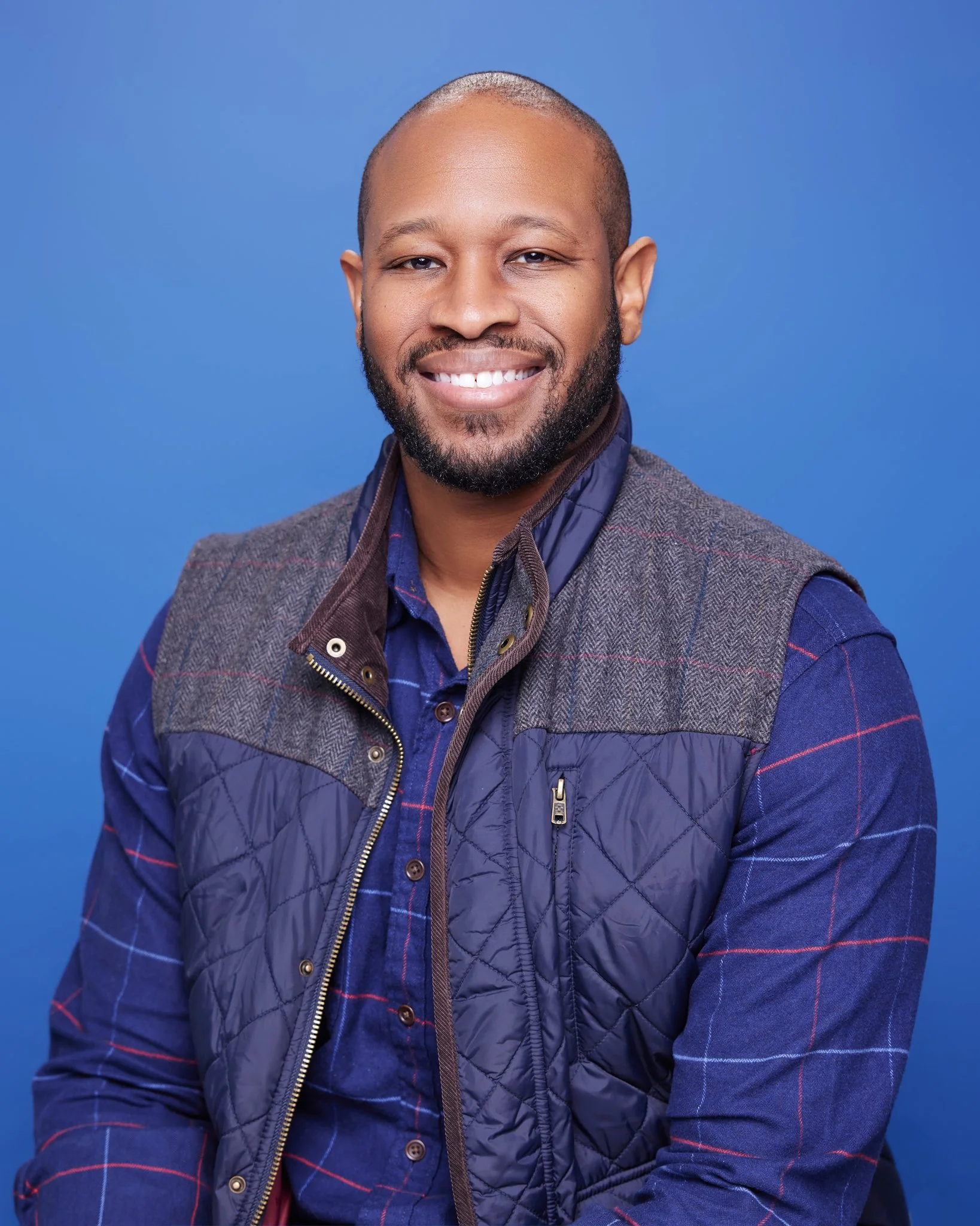 A smiling man with a beard and short hair, wearing a blue checkered shirt and a dark and gray quilted vest, posing against a blue background.