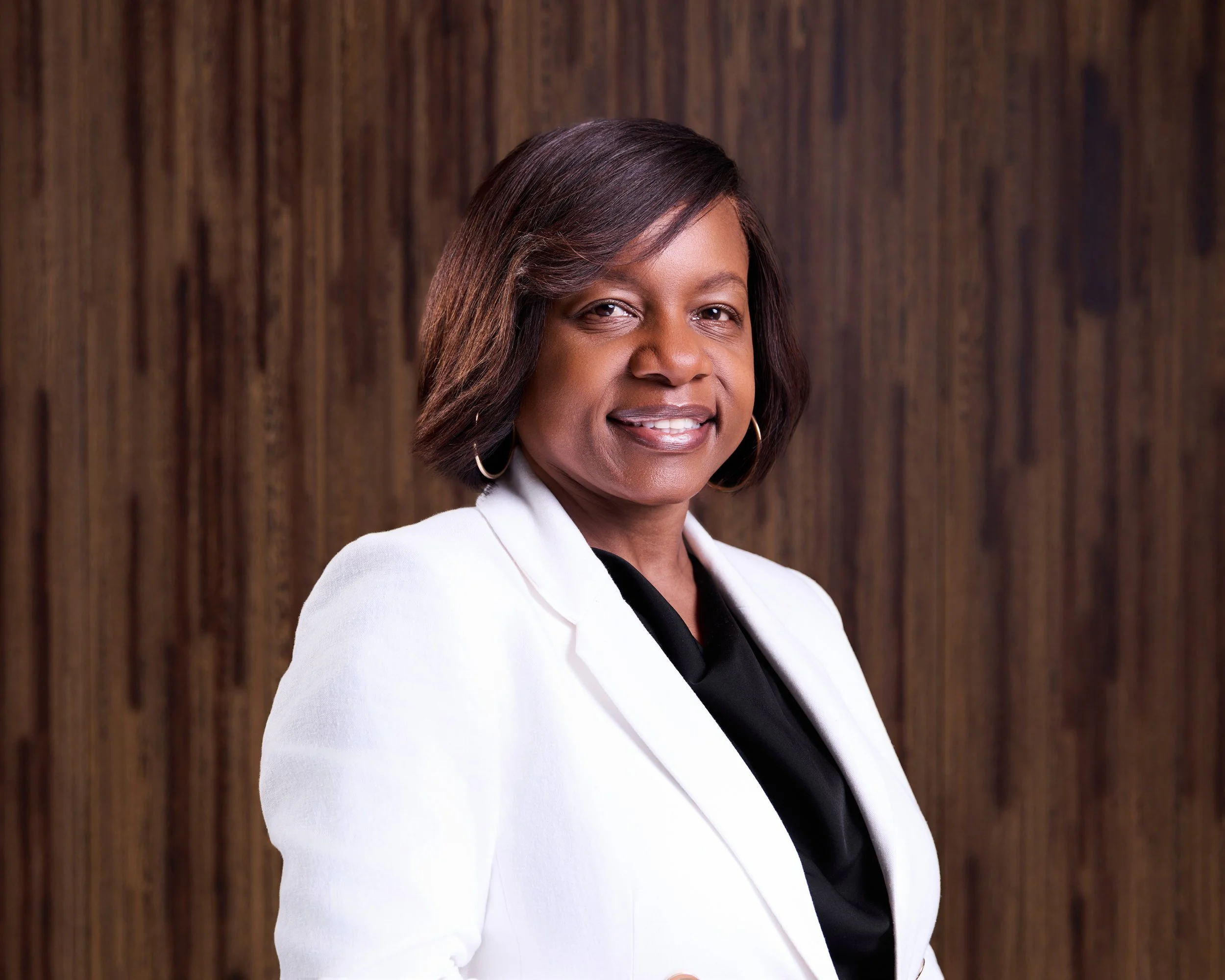 A headshot of a woman with shoulder-length dark hair, wearing a white blazer and a black top, standing in front of a wood-paneled wall and looking at the camera.