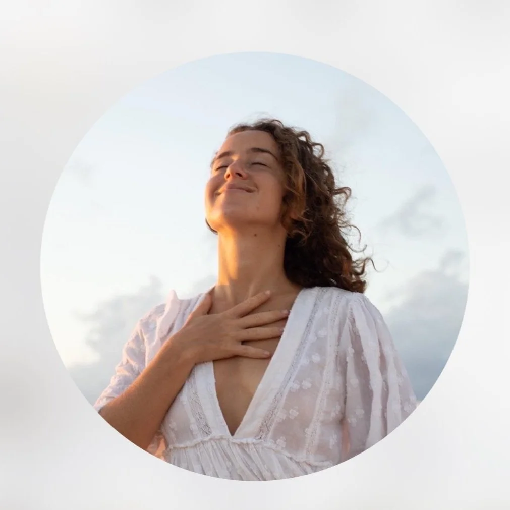 A woman with curly hair in a white blouse, smiling with eyes closed and hand on her chest, outdoors during the day with a cloudy sky.