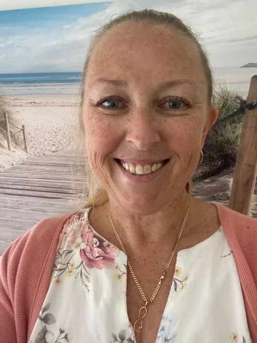 A smiling woman wearing a floral blouse and a pink cardigan stands in front of a beach-themed background with a wooden pathway leading to the ocean.