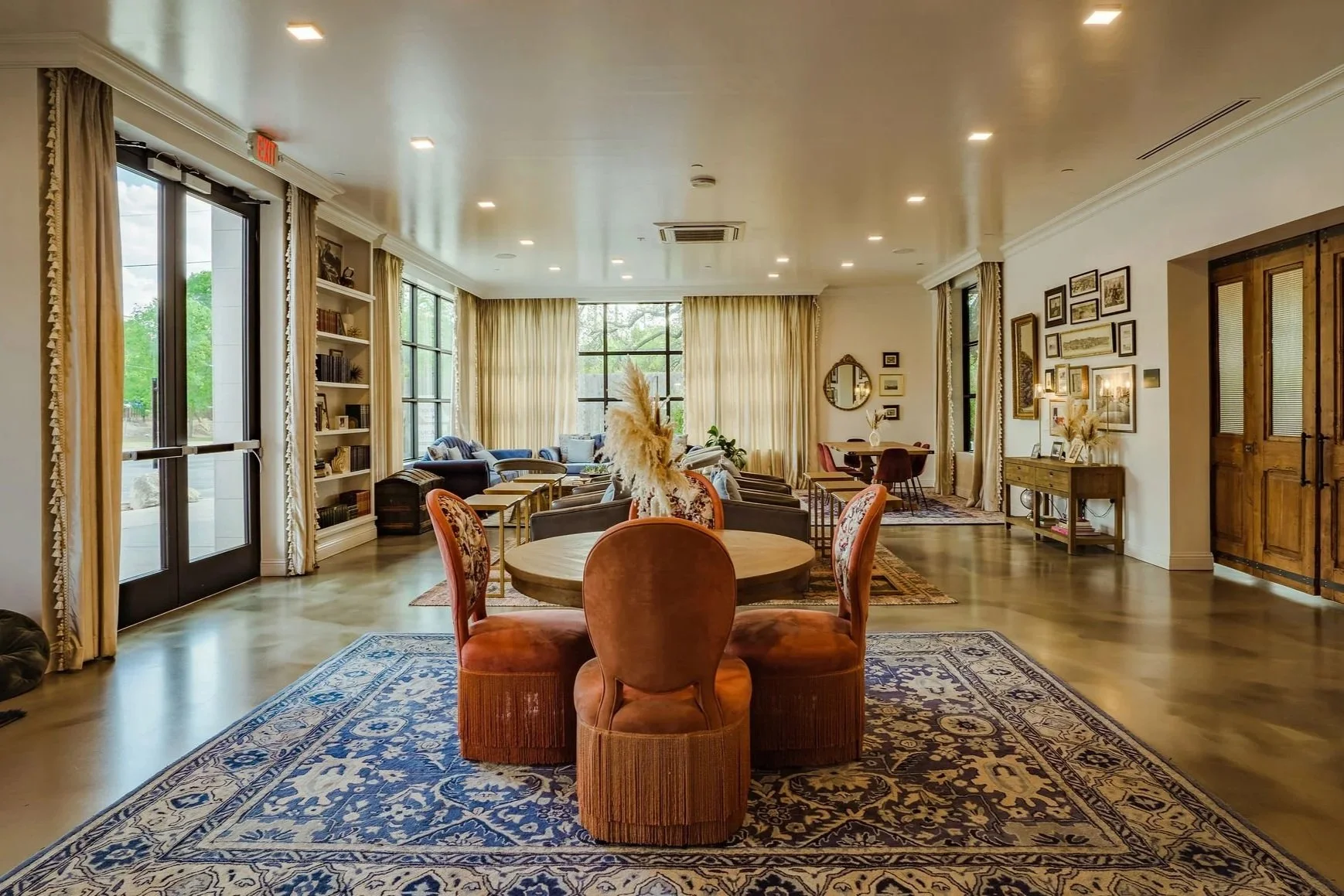 Spacious living room with round dining table, vintage-style chairs, and cozy seating area in the background, illuminated by natural light from large windows and glass doors, decorated with framed pictures and plants.