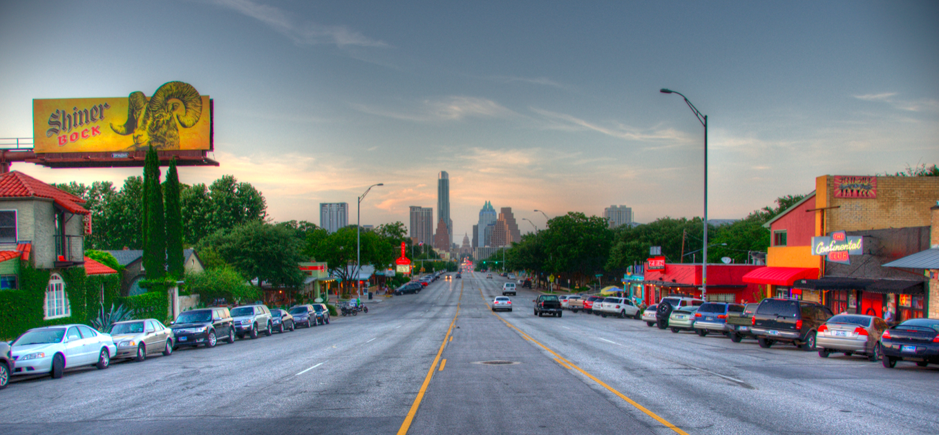 City street view with parked cars on both sides, trees, buildings with colorful signs, and the downtown skyline in the background under a partly cloudy sky at sunset.