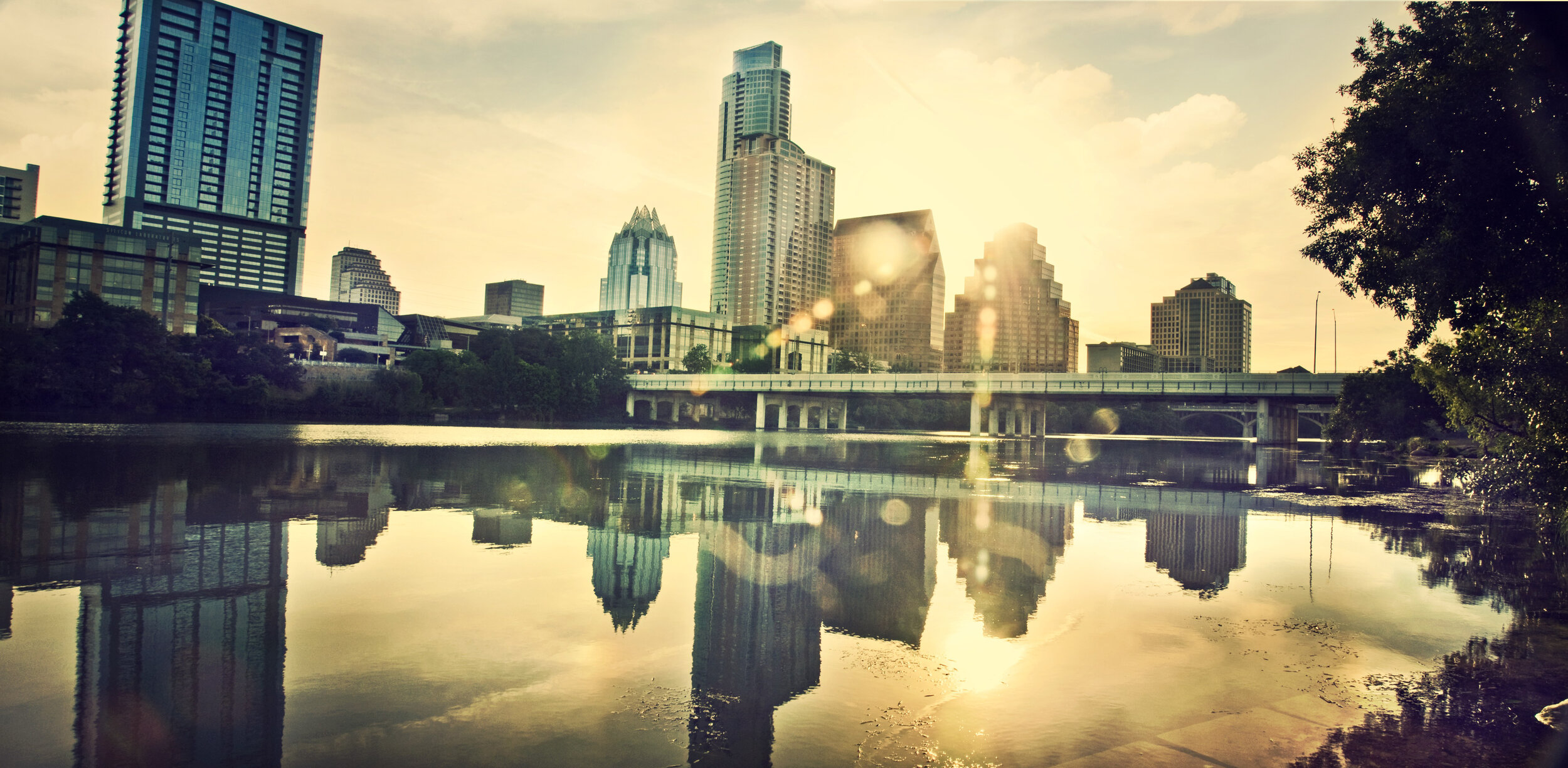 City skyline with tall buildings and a bridge over a river, with the sun setting or rising and creating reflections on the water, surrounded by trees.
