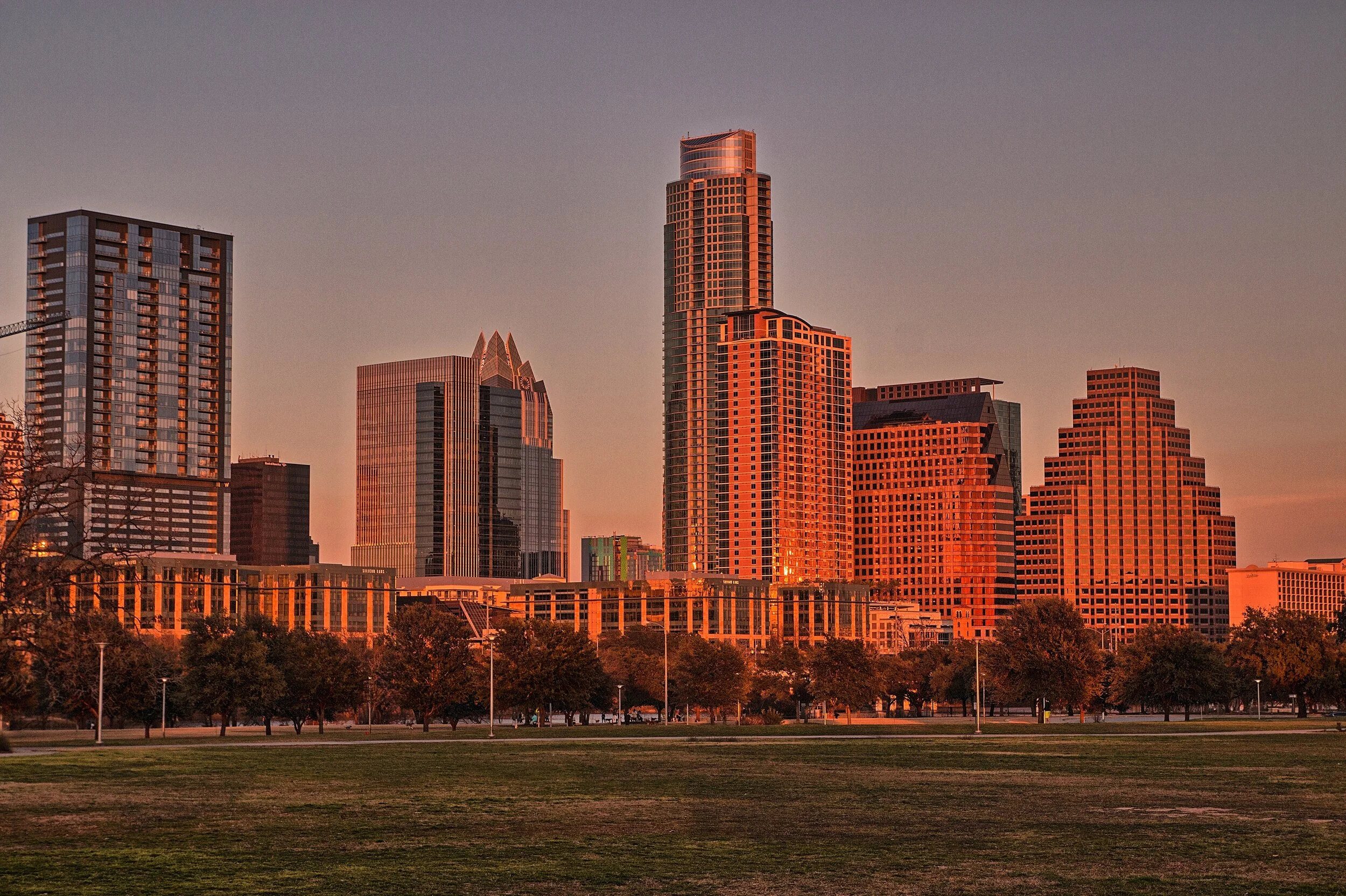 Skyline view of downtown Austin, Texas at sunset, with tall skyscrapers and a park with trees in the foreground.