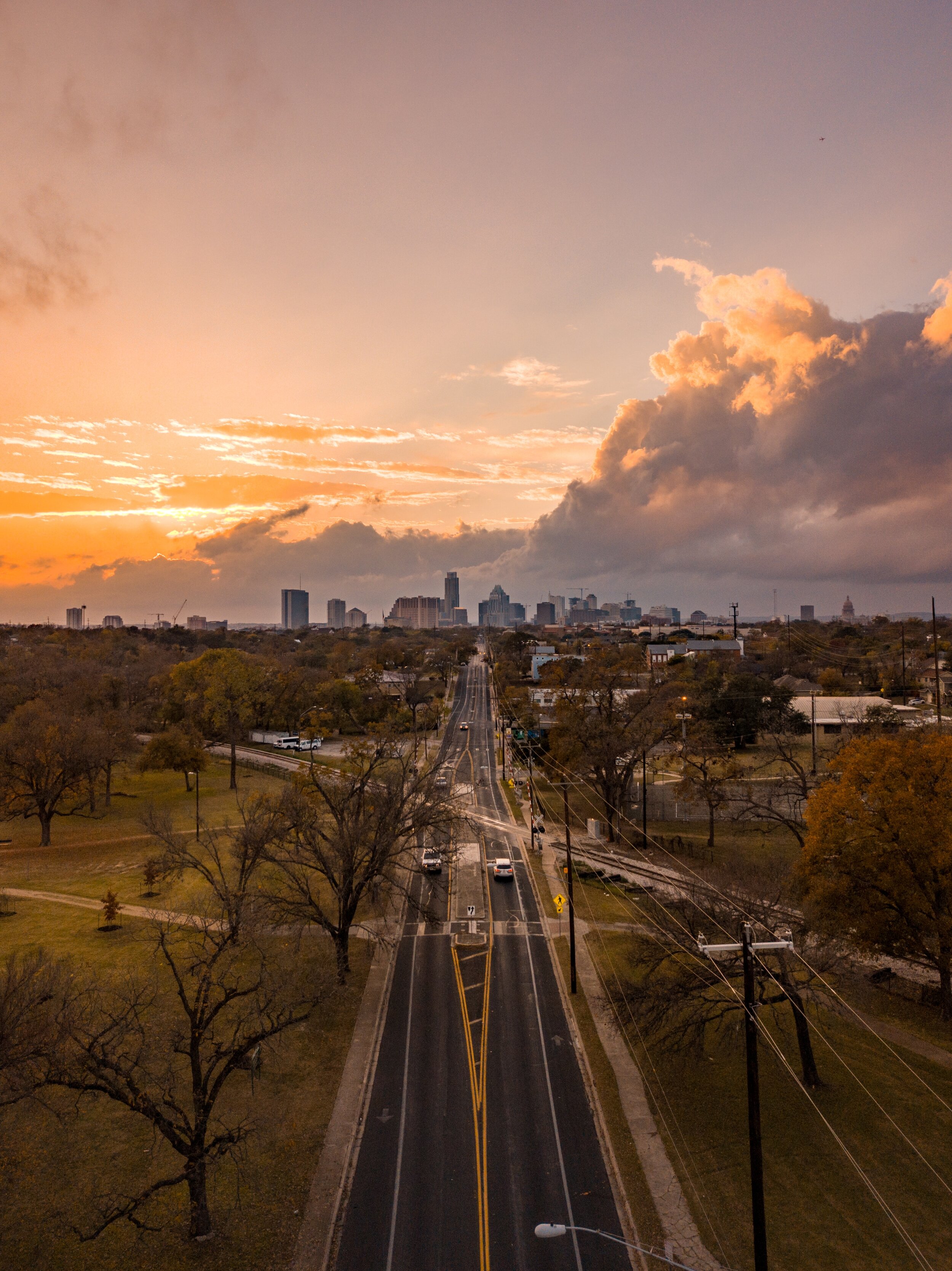 A city skyline at sunset with a long road leading towards tall buildings, trees on both sides, and a partly cloudy sky with orange and purple hues.