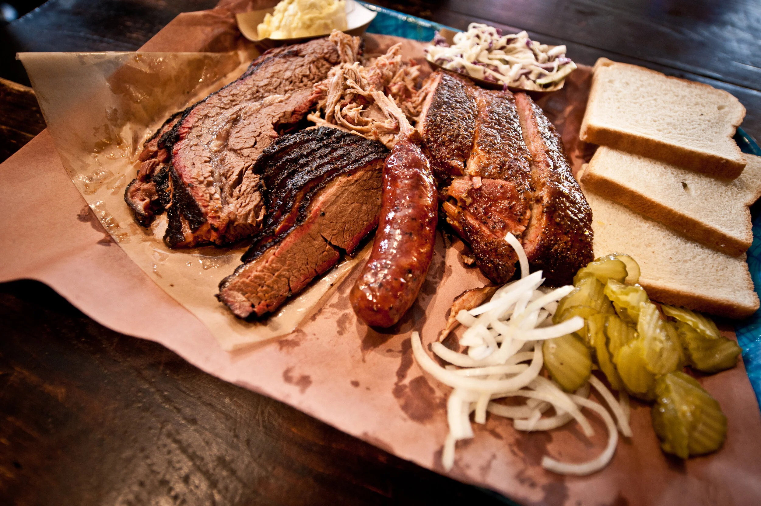 Plate of barbecue meats including brisket, sausage, and ribs, served with sliced white bread, pickles, and coleslaw on a blue tray.