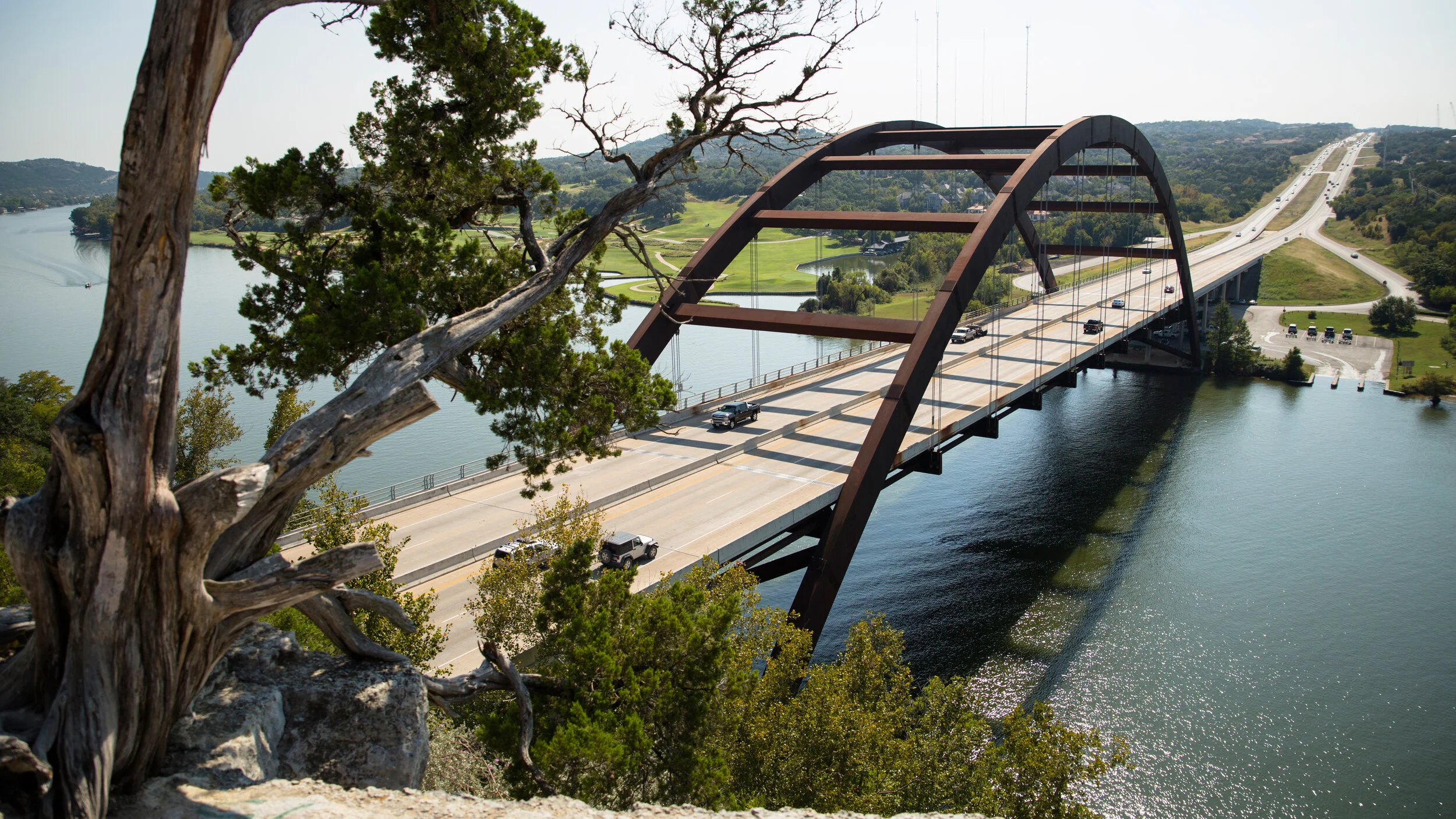 A large steel arch bridge over a river with vehicles driving across it, with trees in the foreground and hills in the background.