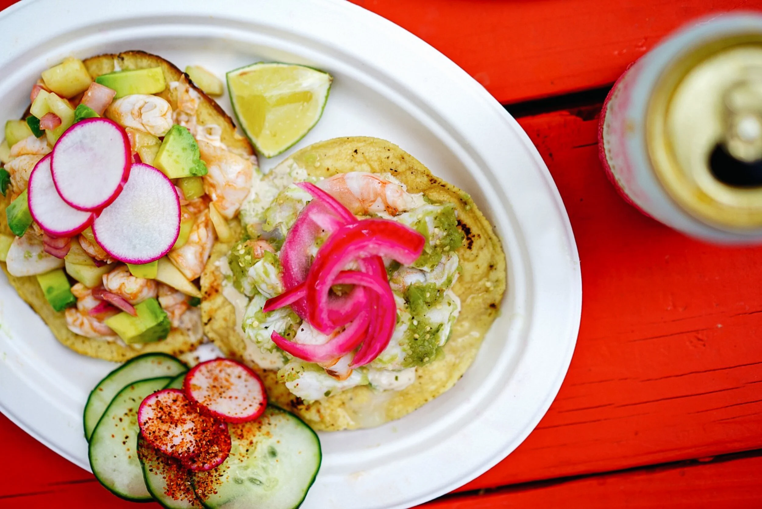 Two shrimp tacos with radish slices, a lime wedge, cucumber slices, and a beer on a red wooden table.