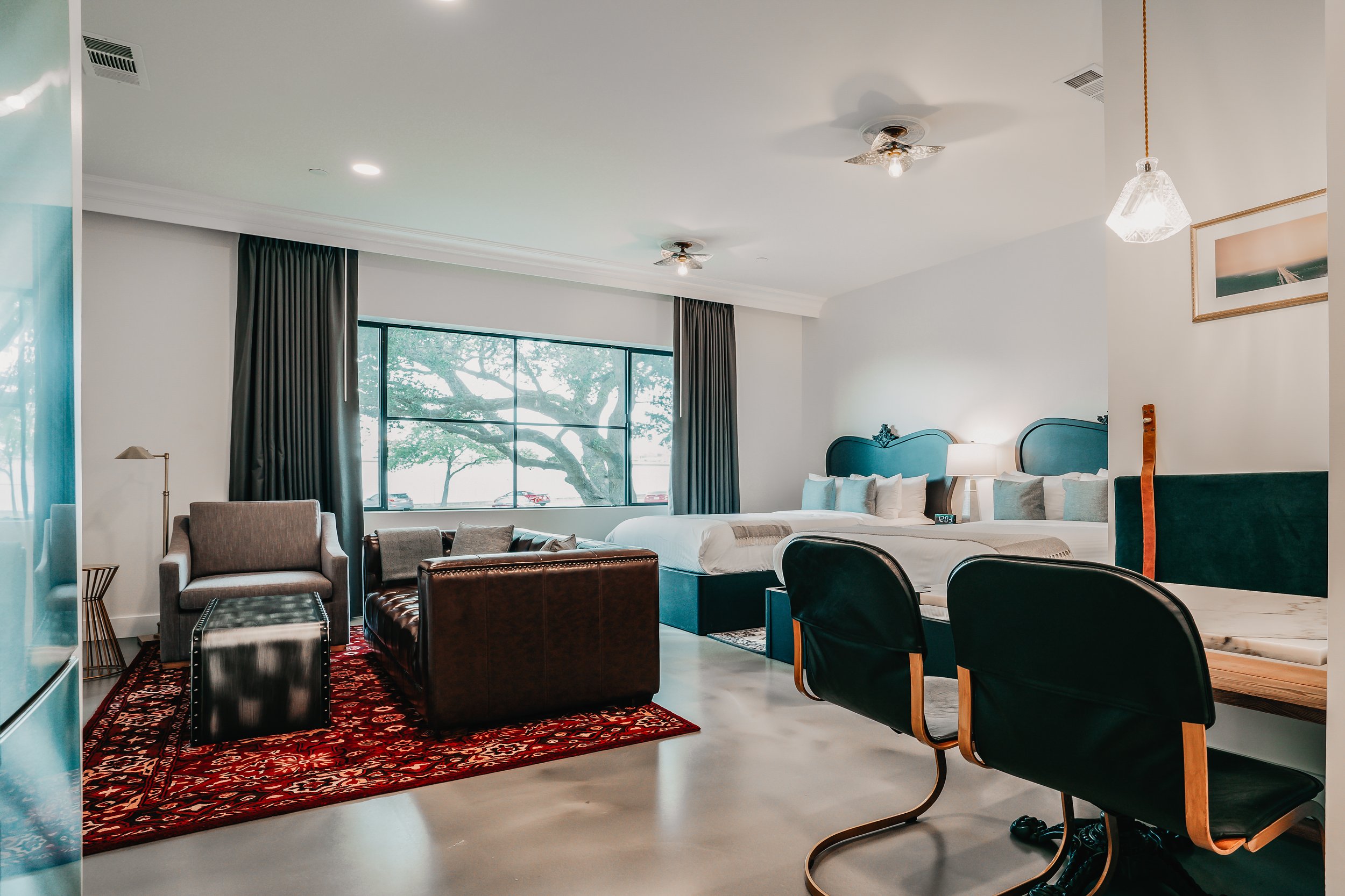 Hotel room with two beds, sitting area, large window showing trees, dark curtains, modern decor, and leather chairs.