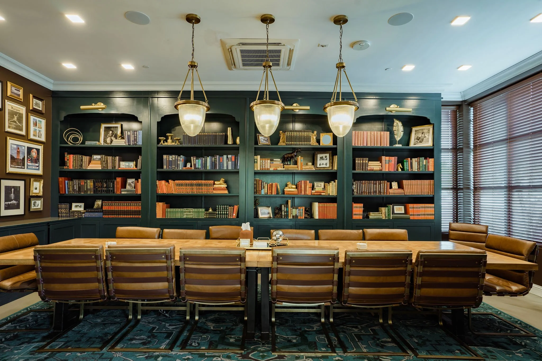 A large conference room with a long wooden table surrounded by brown leather chairs. Behind the table, there is a dark green bookshelf with various books and decorative items. Overhead, three pendant lights hang from the ceiling, and there are blinds covering windows on the right side of the room.
