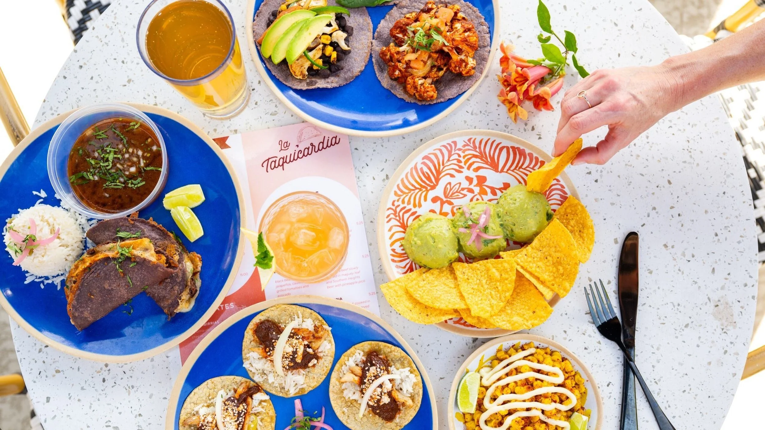 A variety of Mexican dishes on a white table, including tacos, guacamole with tortilla chips, rice, a bowl of enchilada, and small tacos, with a hand reaching for the guacamole and tortilla chips.