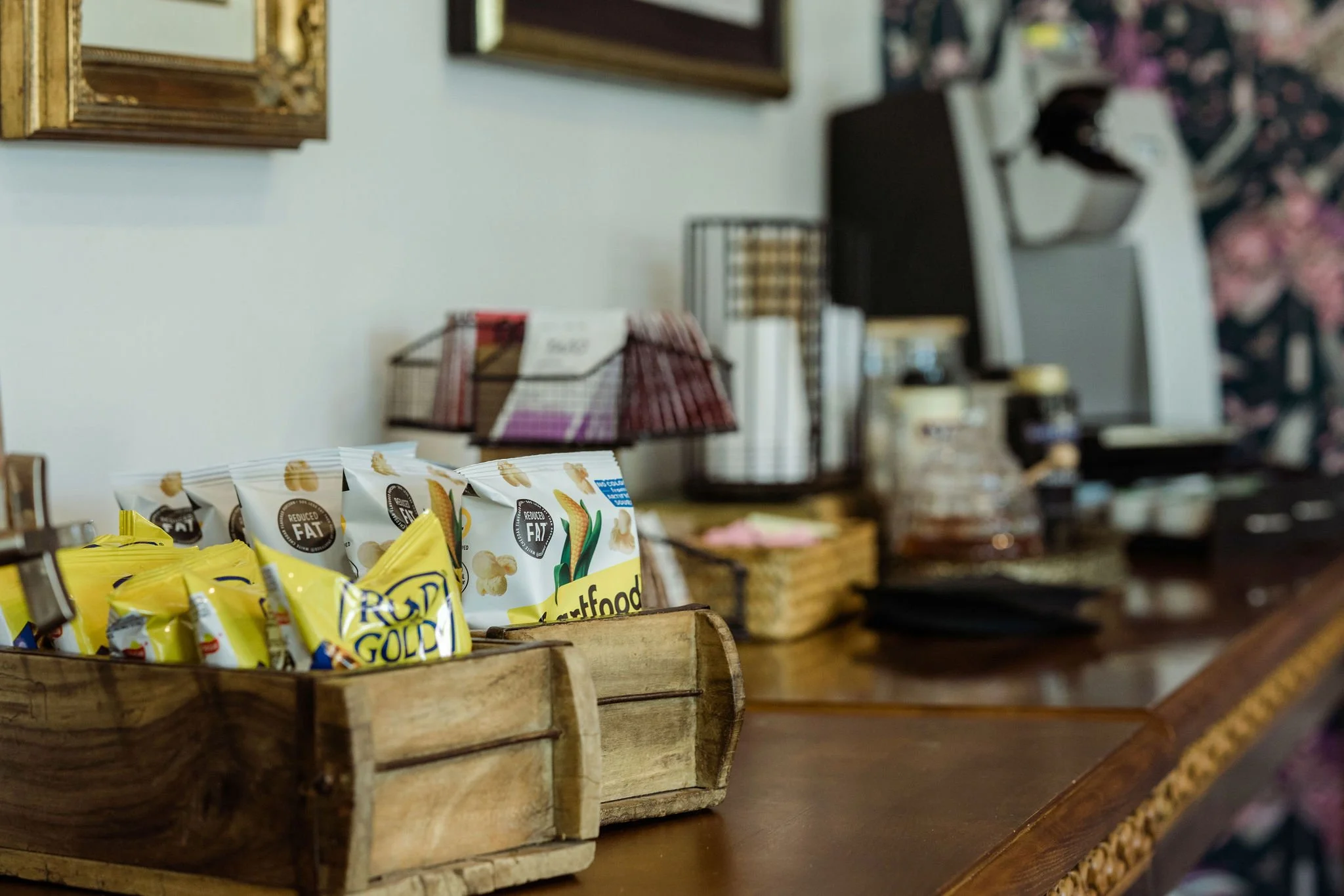 Snack items in wooden baskets on a counter in a cozy cafe or store, with shelves and framed art on the wall in the background.