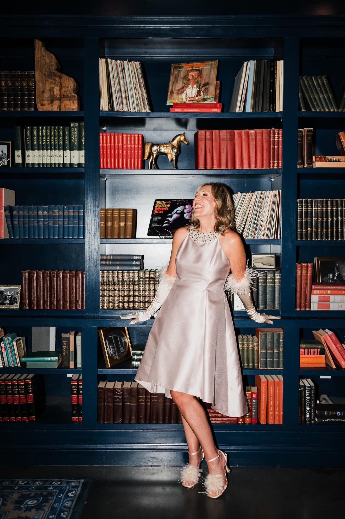 A woman in an elegant dress standing in front of a blue bookshelf, smiling and looking to the side.