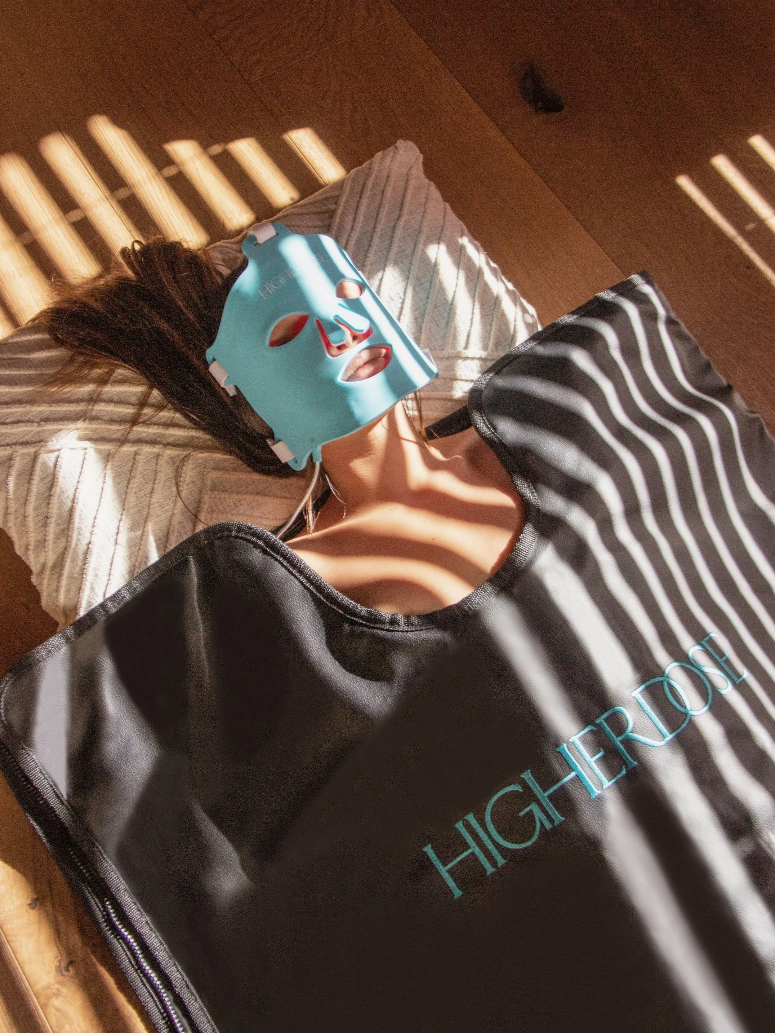Person lying on a bed with sunlight casting shadows, wearing a blue facial mask, covered with a black and white striped blanket, with a HighロRose skincare package nearby.